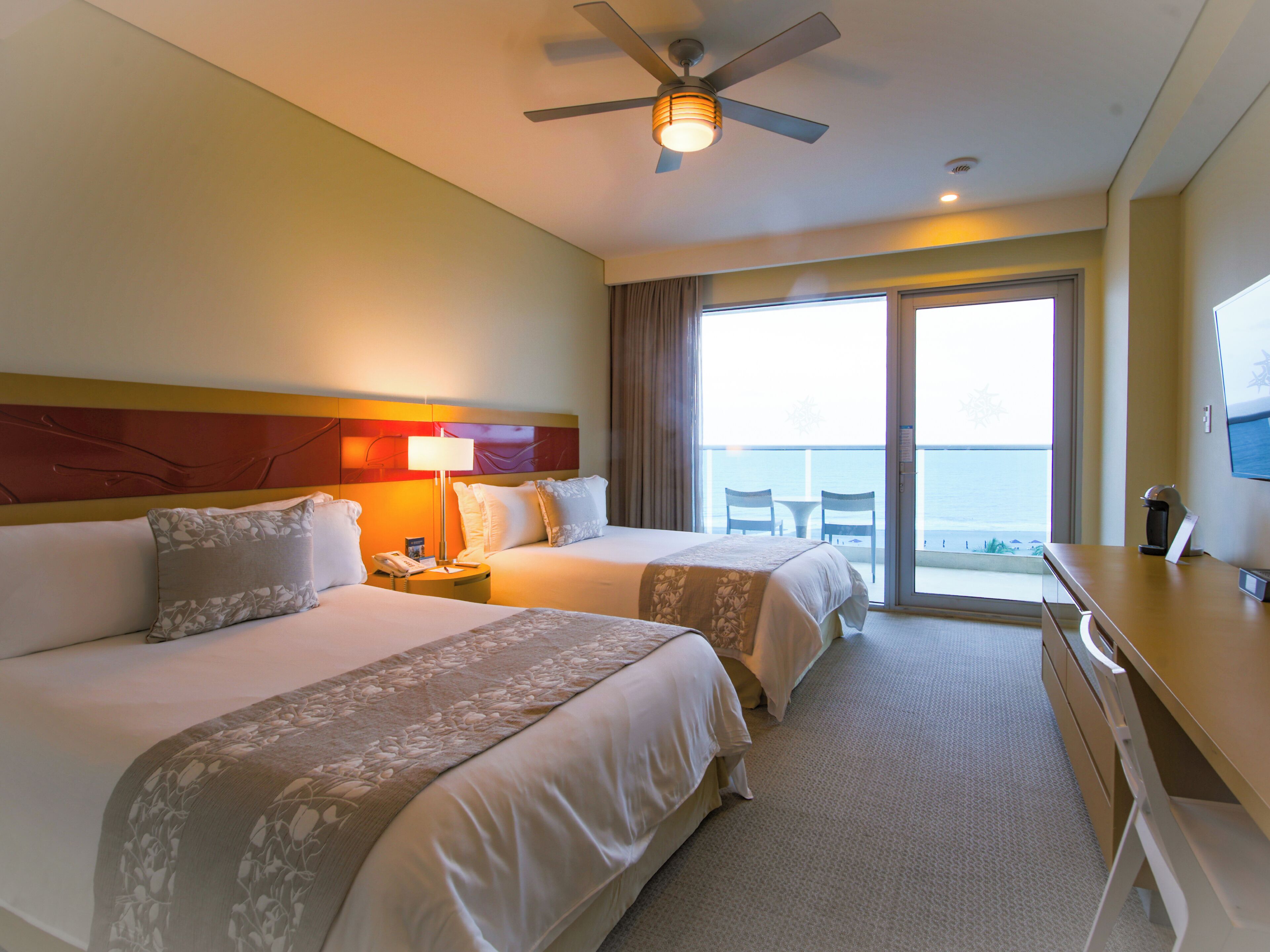 Ein Hotelzimmer mit zwei Betten, einem Schreibtisch, einem Deckenventilator und Blick auf das Meer durch die Glasdoor.