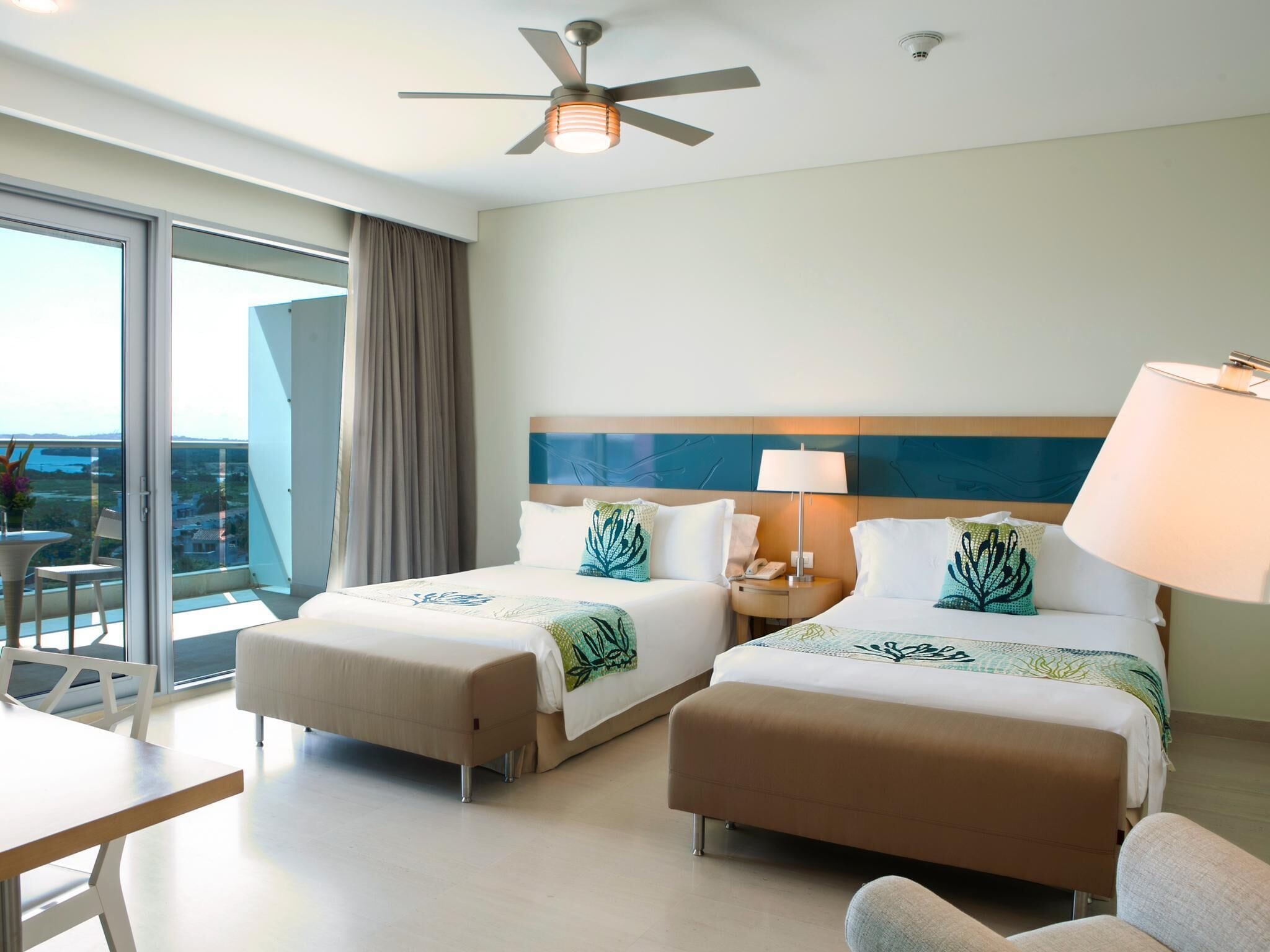 A hotel room with two beds, a ceiling fan, a table, chairs, and a view of the outdoors.