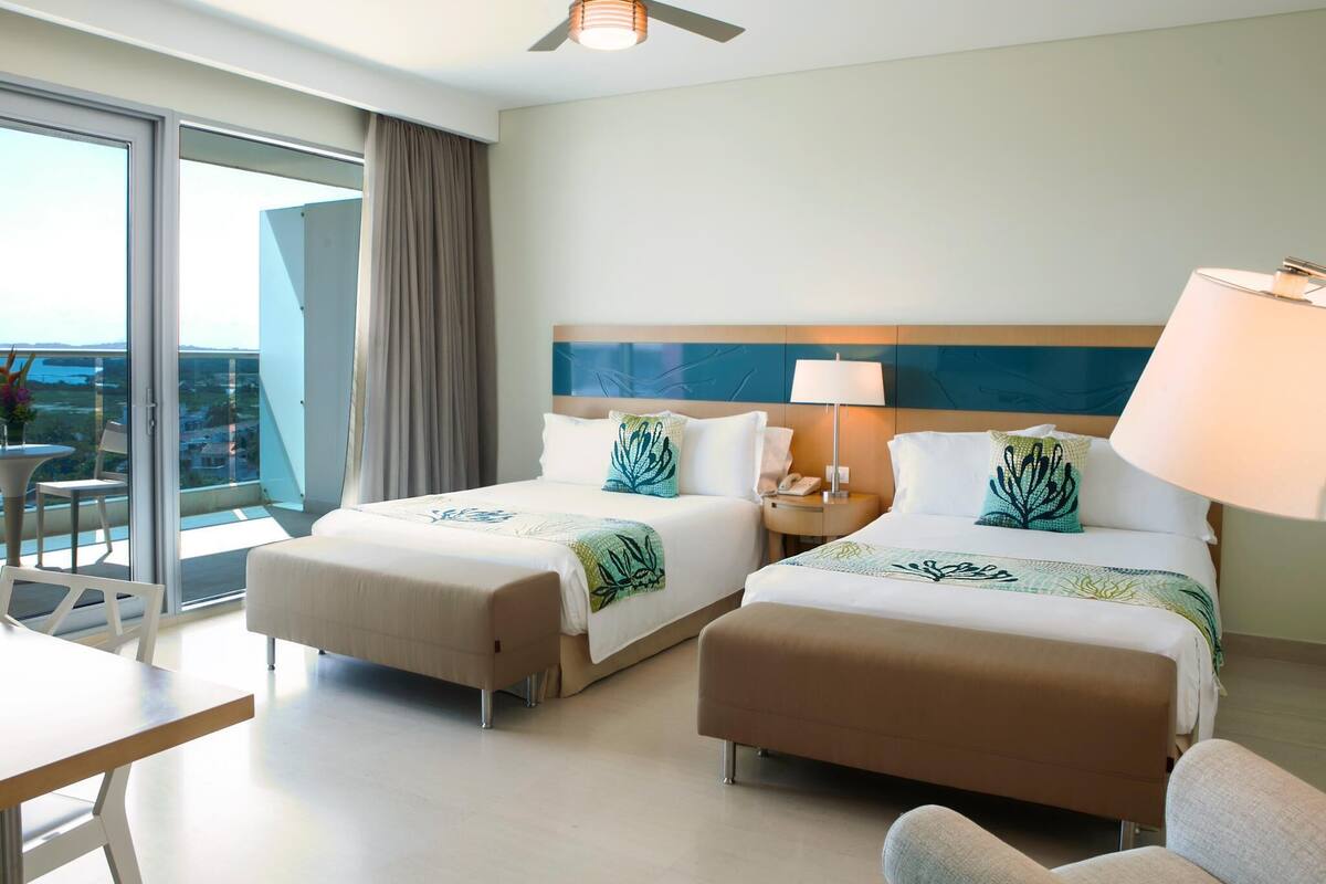 A hotel room with two beds, a ceiling fan, a table, chairs, and a view of the outdoors.