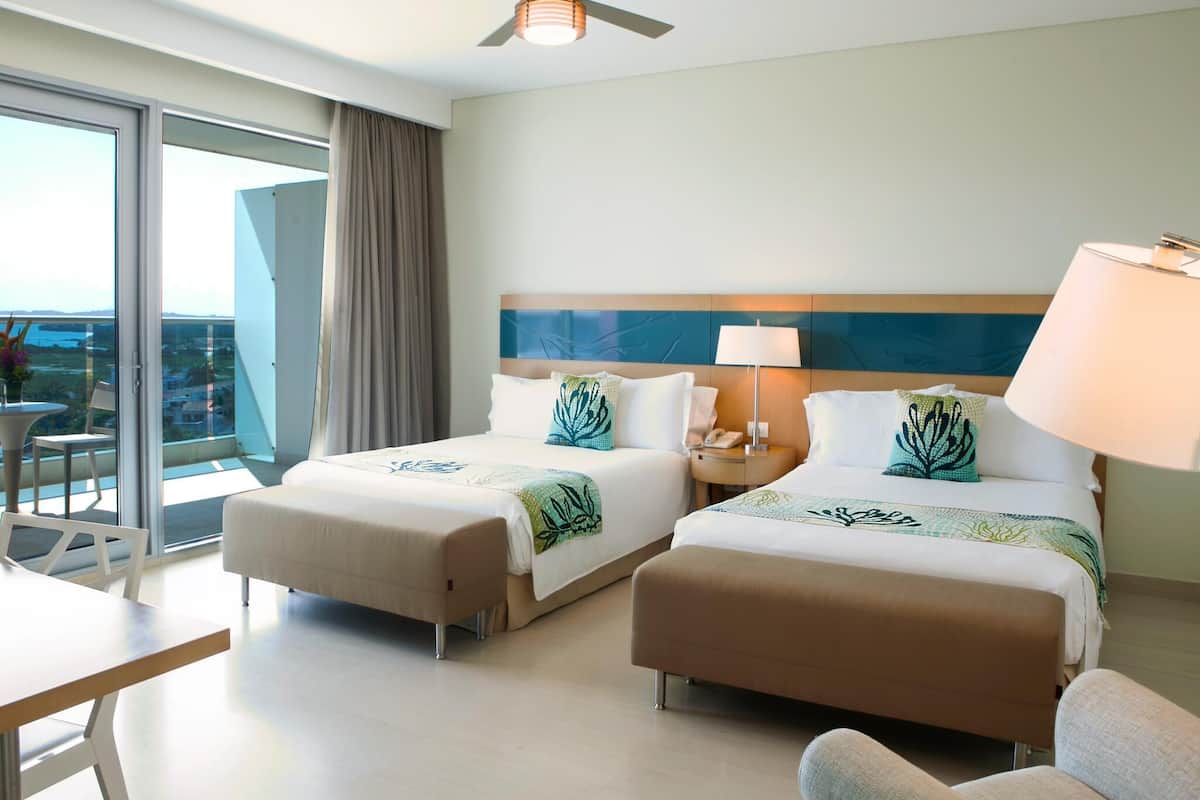 A hotel room with two beds, a ceiling fan, a table, chairs, and a view of the outdoors.