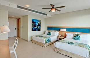 Habitación de hotel con dos camas, ventilador de techo, un cuadro en la pared y un escritorio con una silla.