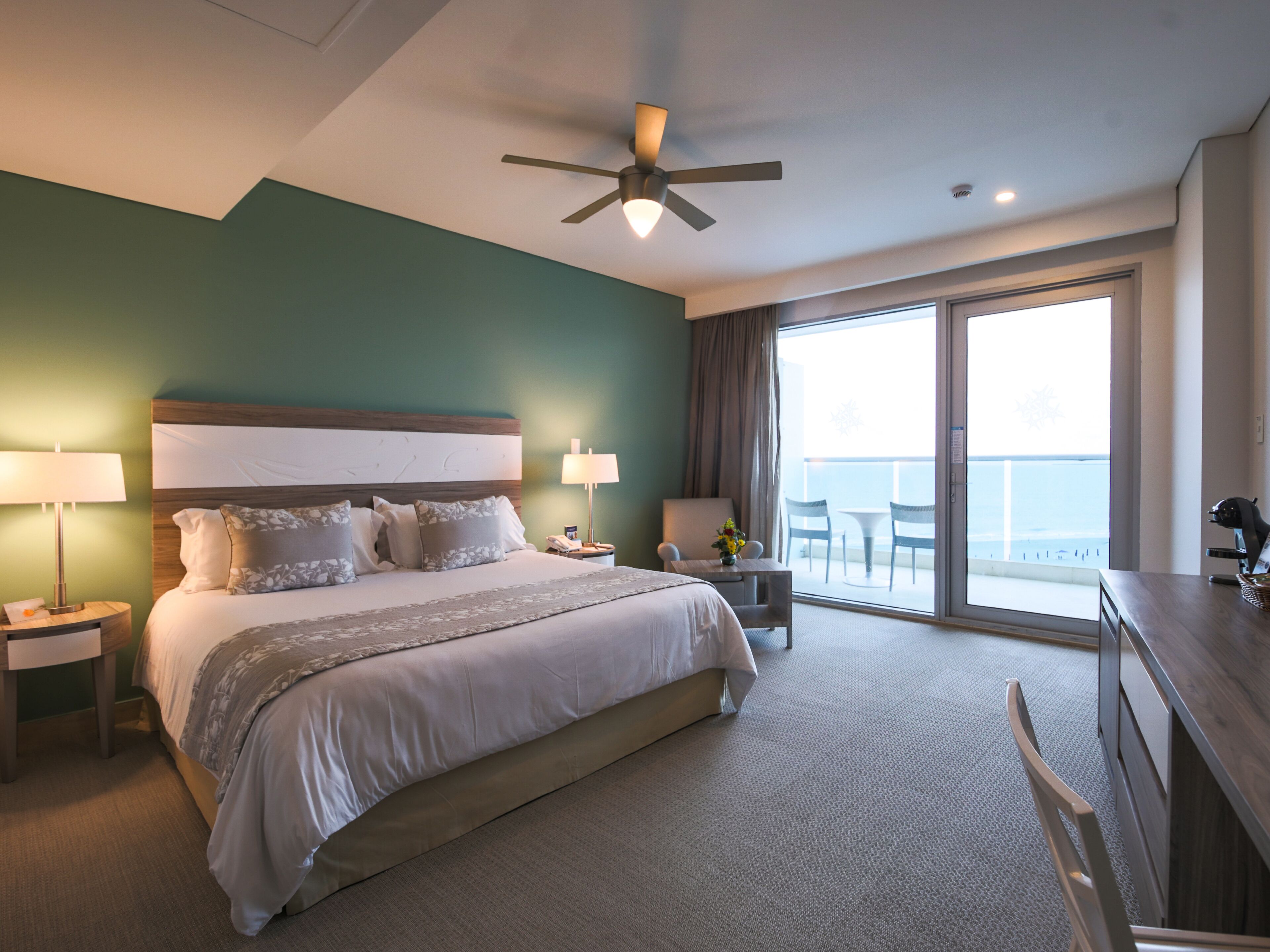 A hotel room with a large bed, two bedside tables, a desk, a chair, and a balcony with a view.