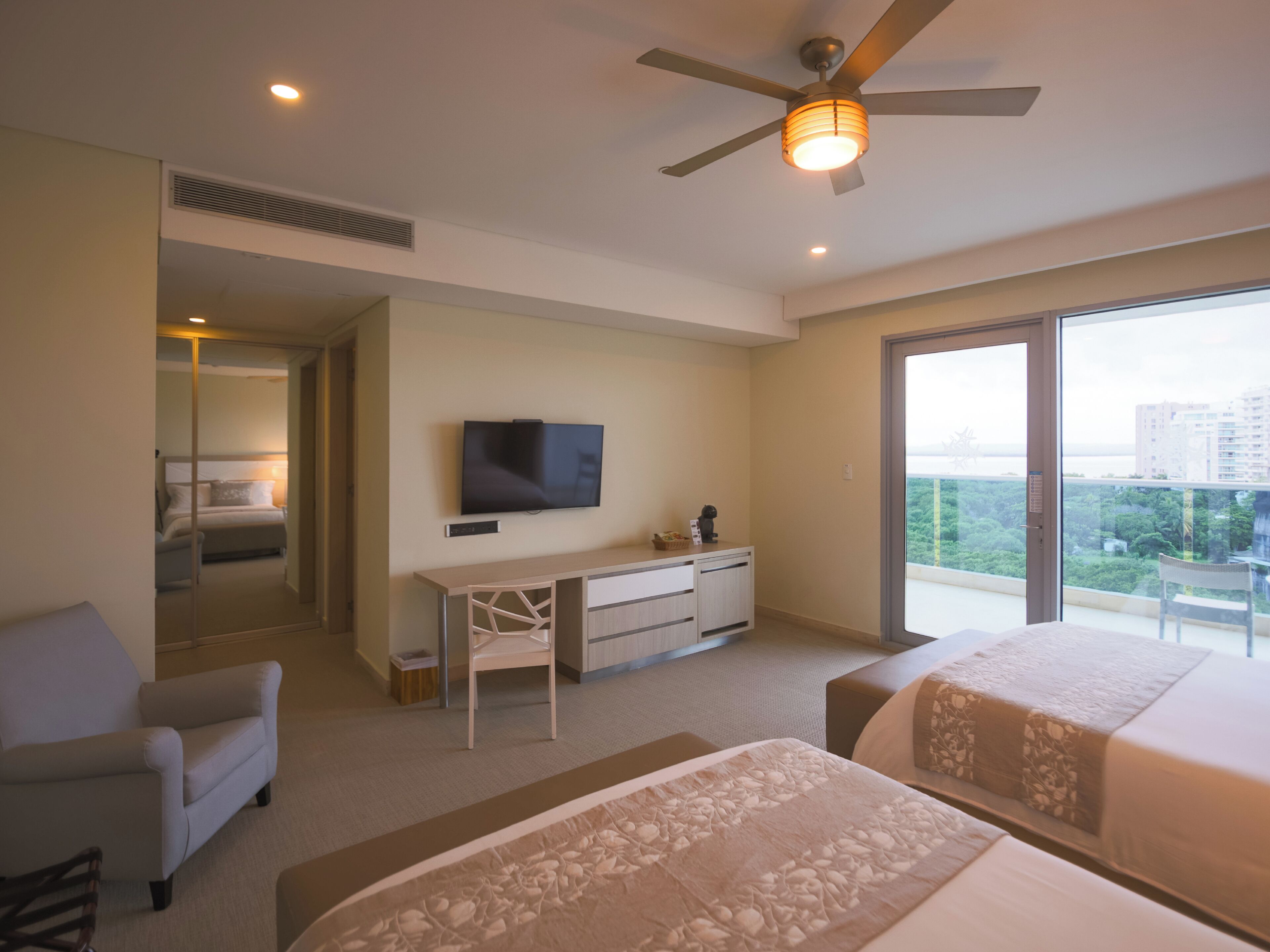 A modern hotel room with a large bed, a desk with a flat-screen TV, a chair, and a balcony with a city view.