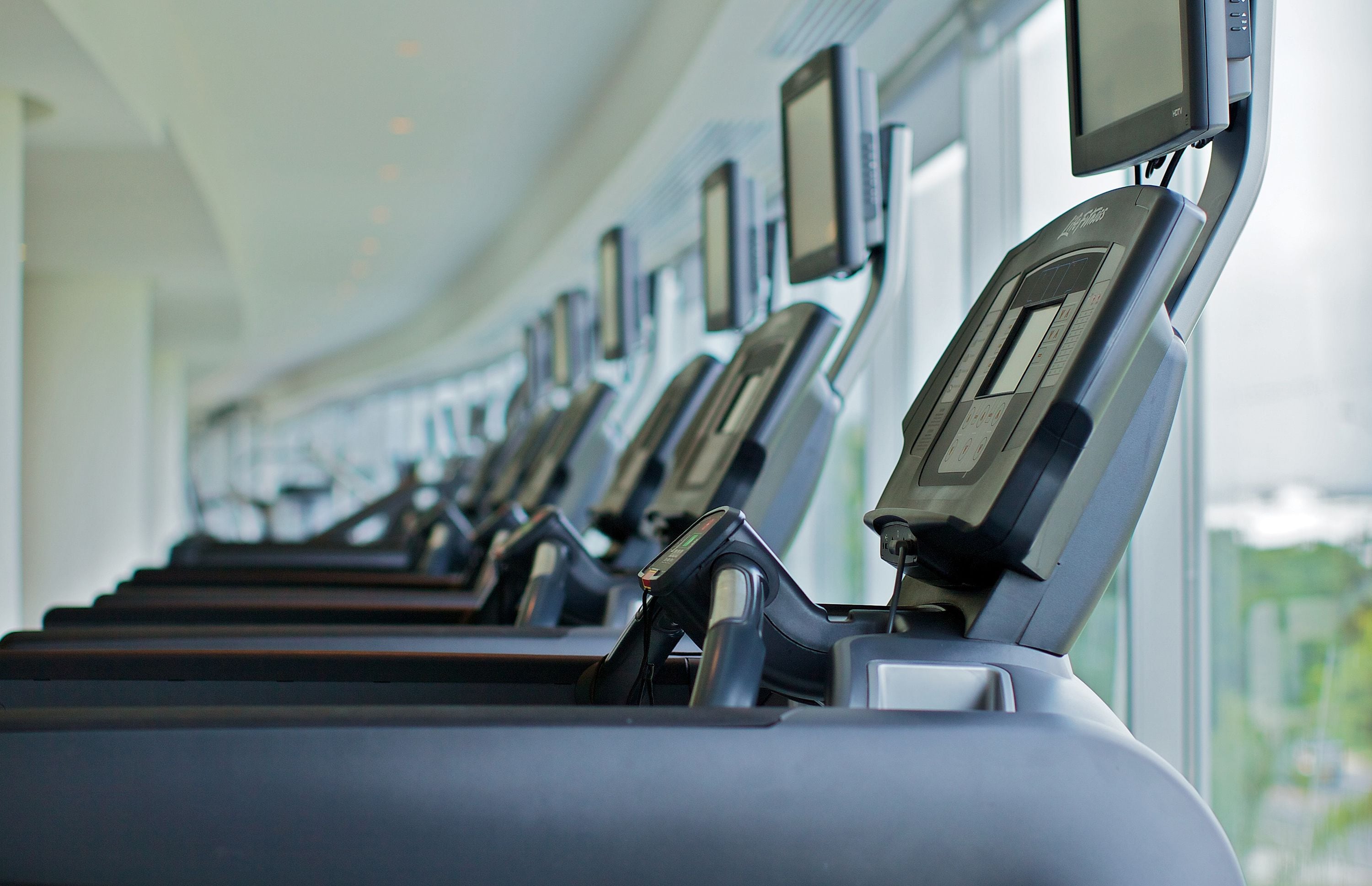 A row of stationary bikes with digital displays, arranged in a gym setting with large windows.
