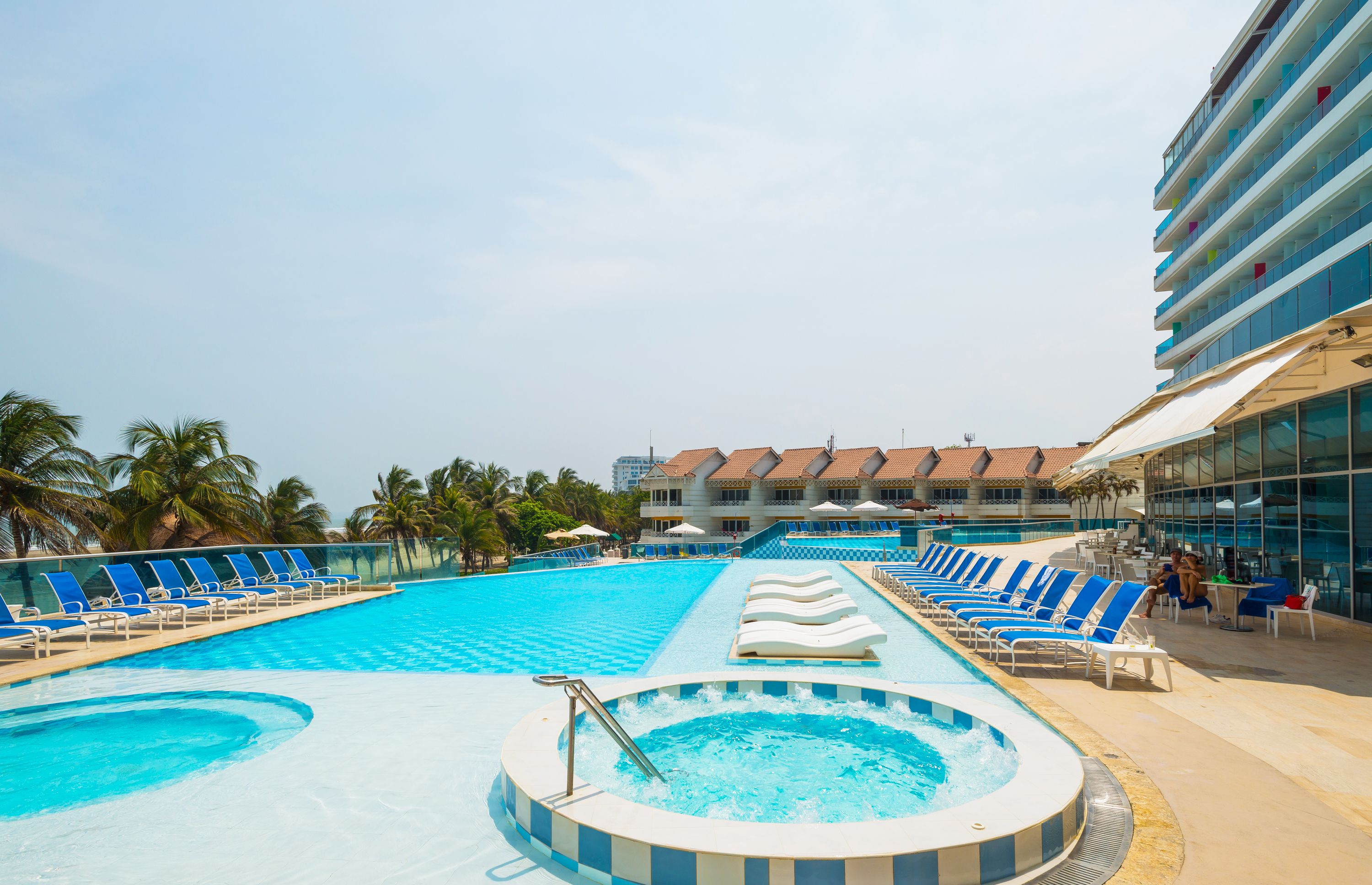 A swimming pool with lounge chairs, a hot tub, and a building with a tiled roof in the background.