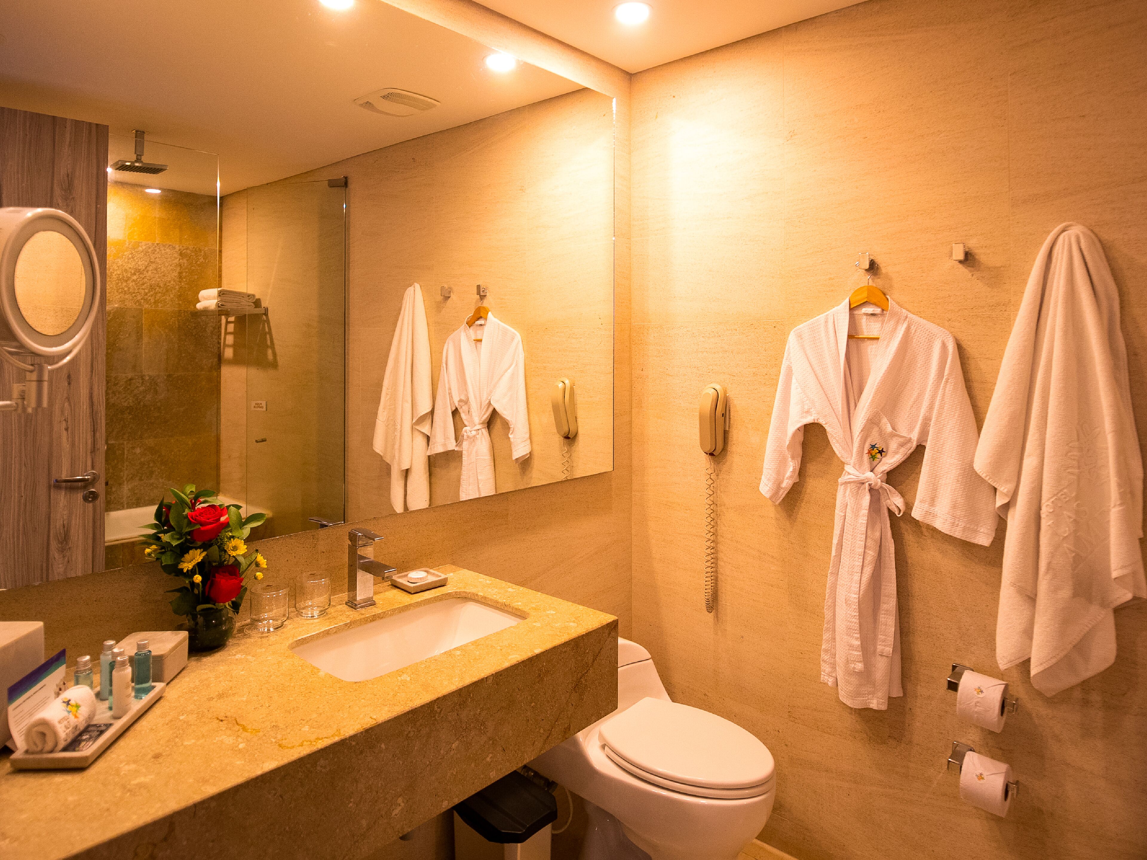 A modern bathroom with a marble sink, a toilet, and a shower. There are white towels and robes hanging on hooks, and a phone on the wall.