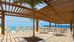 A covered outdoor seating area with wooden tables and chairs, a palm tree, and a view of the ocean.