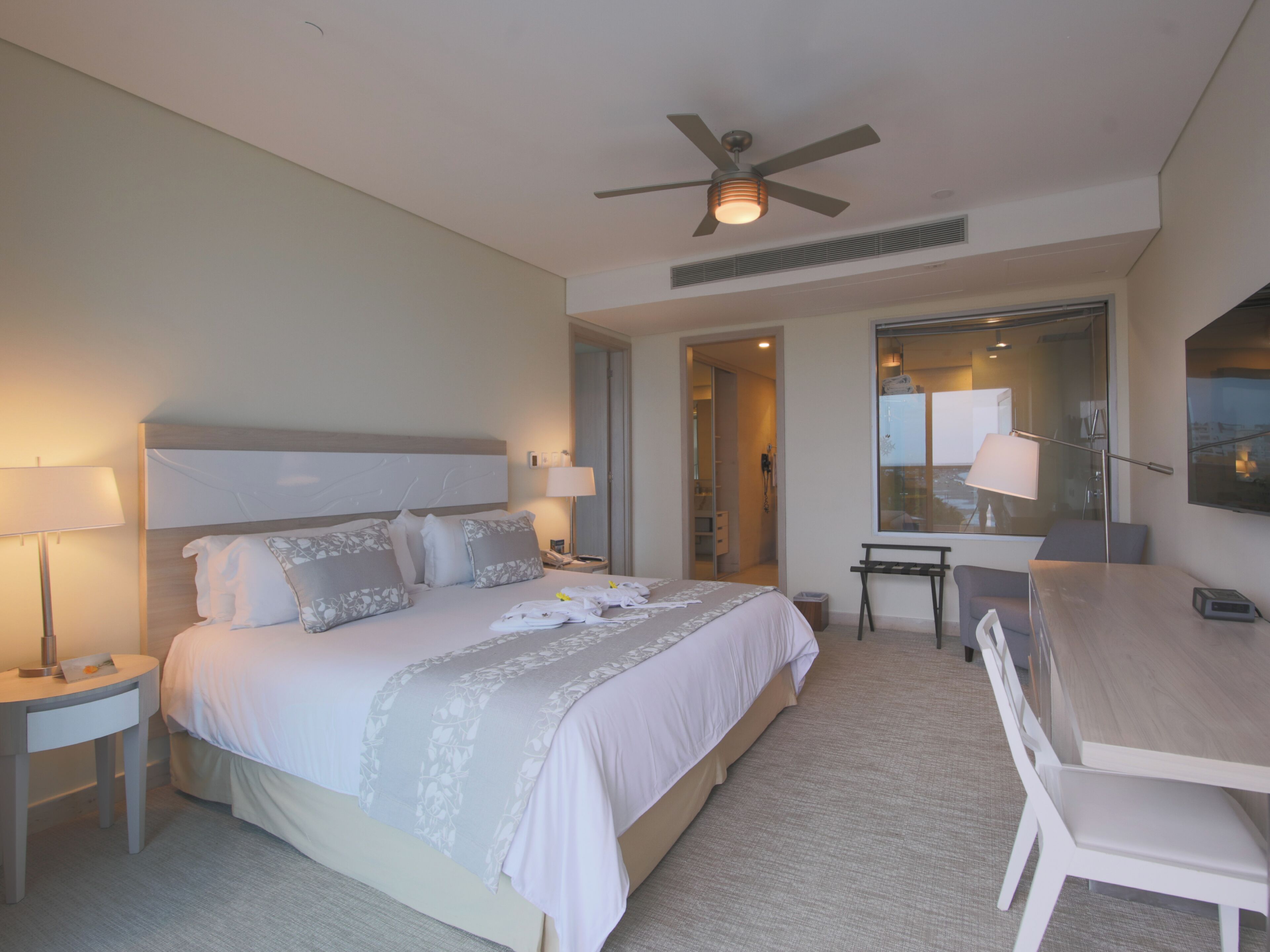 A hotel room with a large bed, bedside tables, a desk, and a chair. There is a ceiling fan and a window with a view of the city.