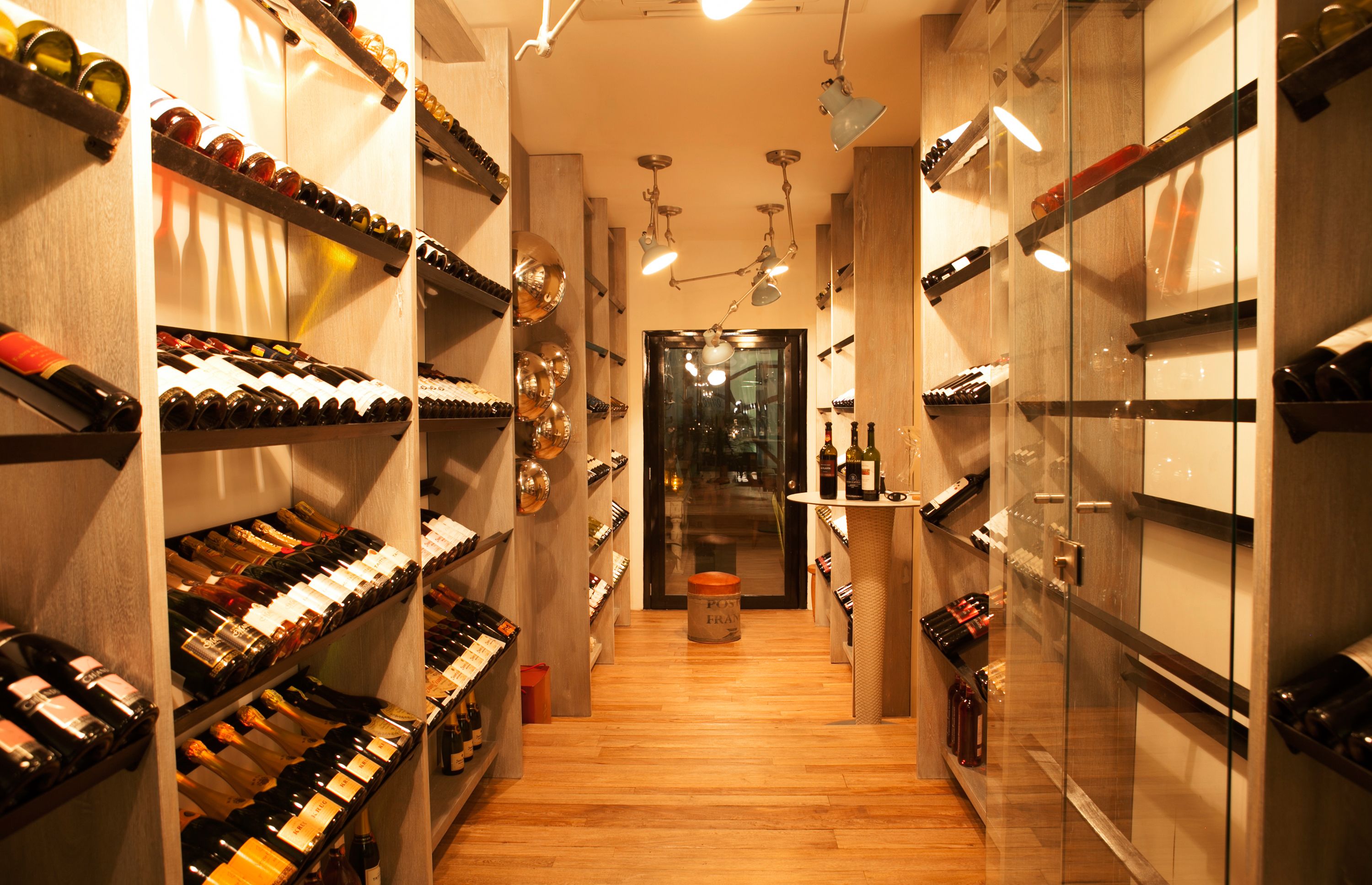 A wine cellar with wooden shelves and hanging wine racks, a central wooden floor, and a glass door leading to another room.