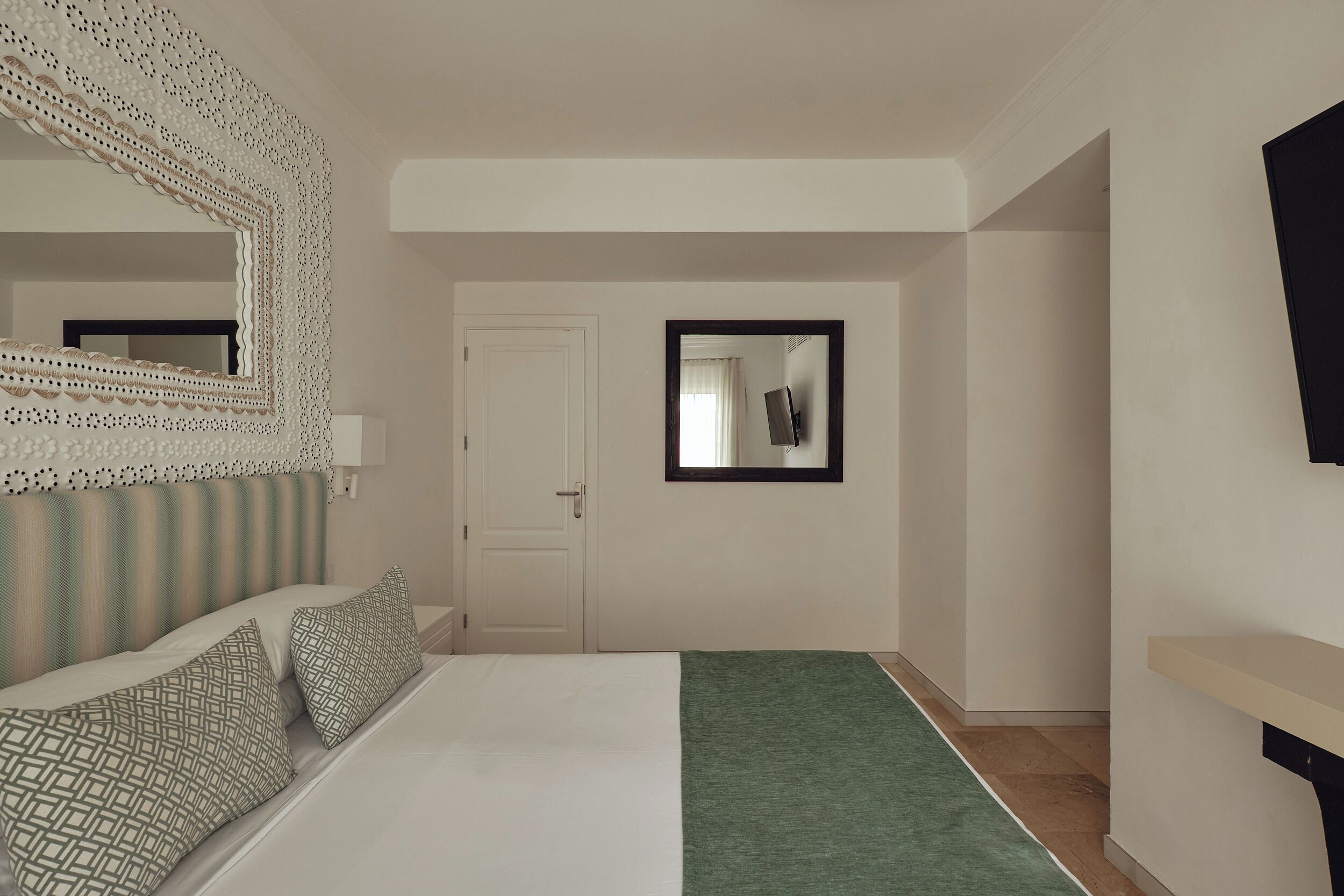 A bedroom with a bed, a green throw blanket, a decorative mirror, and a door.