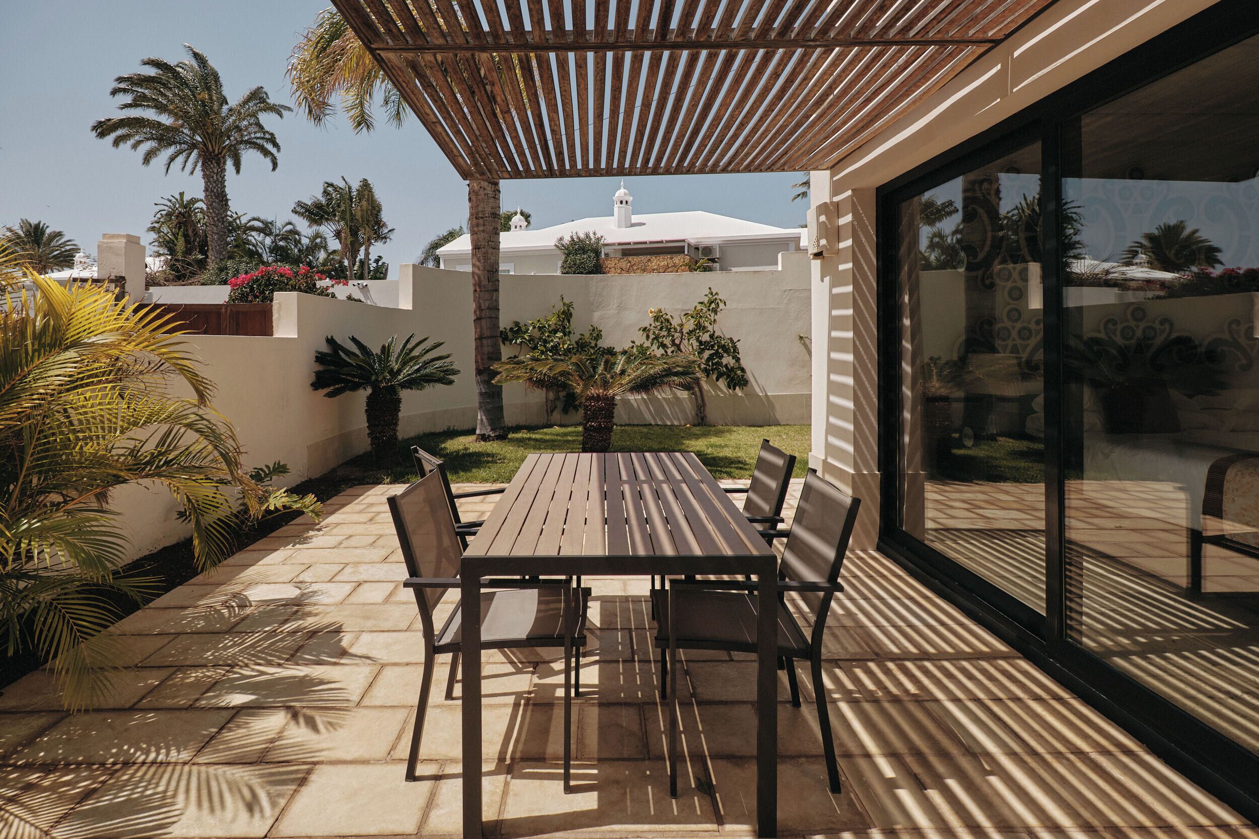 An outdoor patio with a wooden table and chairs, surrounded by palm trees and potted plants.