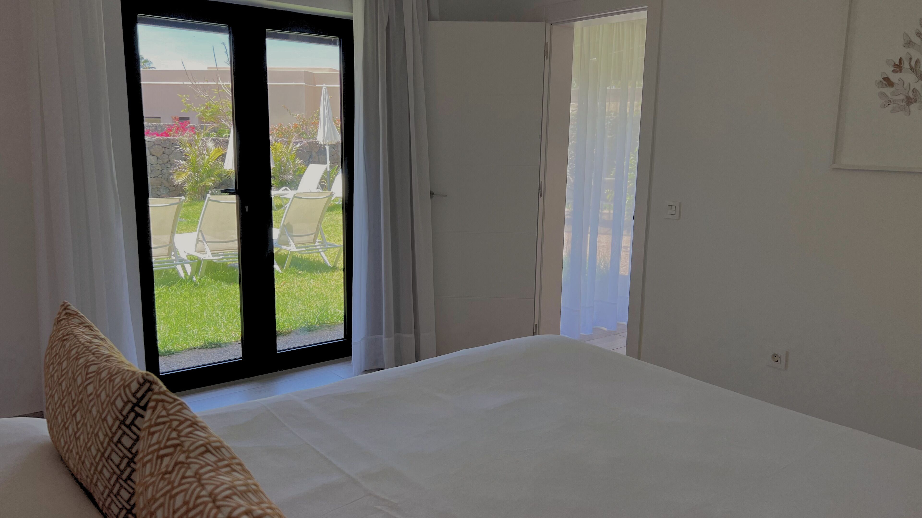 Ein Schlafzimmer mit einem Bett, einem großen Fenster und Blick auf einen Außenbereich mit Stühlen und Pflanzen.