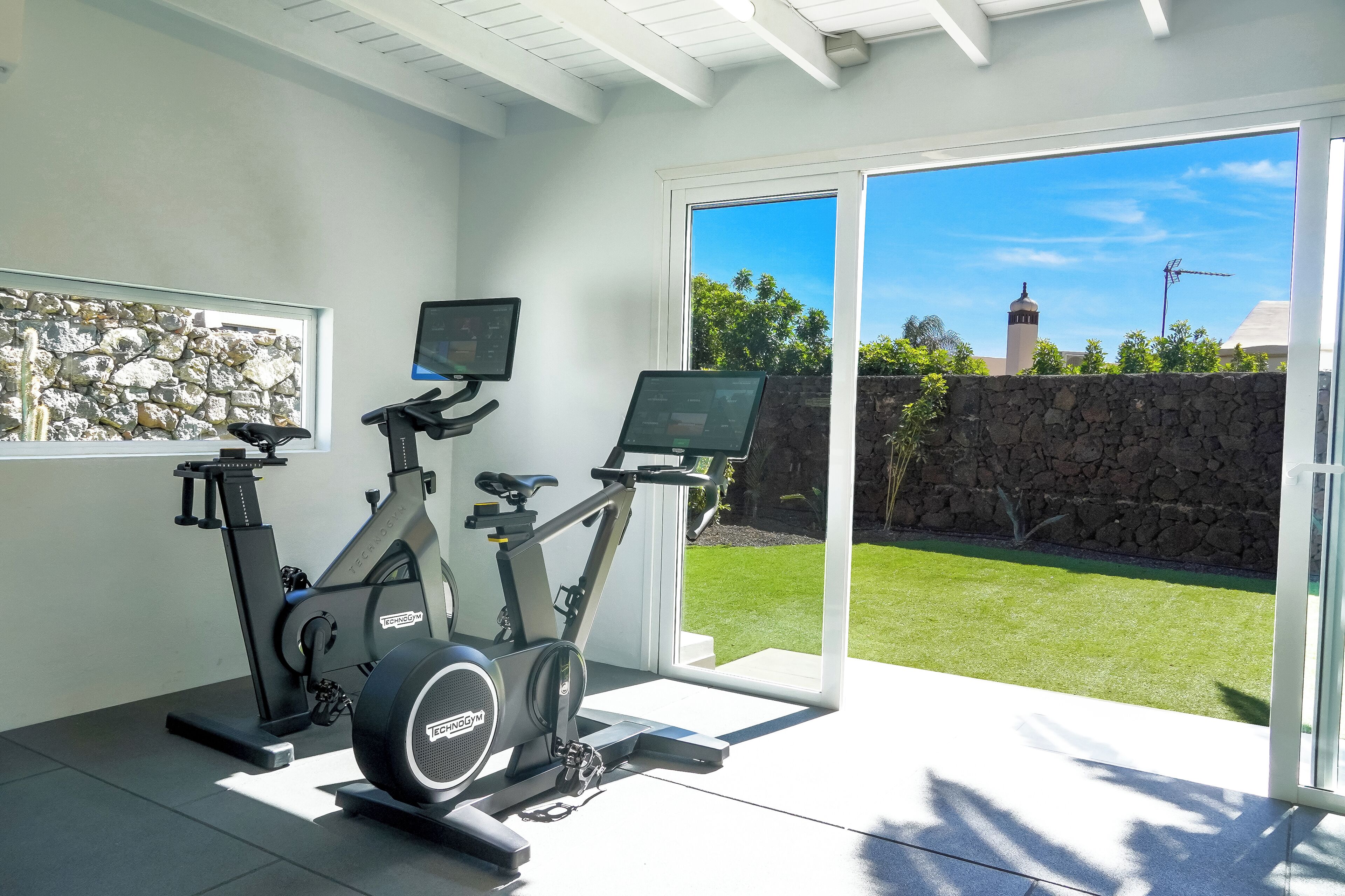 A home gym with stationary bikes and a treadmill, a stone wall, and a large sliding glass door leading to a lawn.