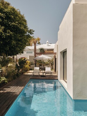 Una piscina con cubierta de madera, rodeada de abundante vegetación y un edificio blanco con fachada blanca y una pequeña torre.