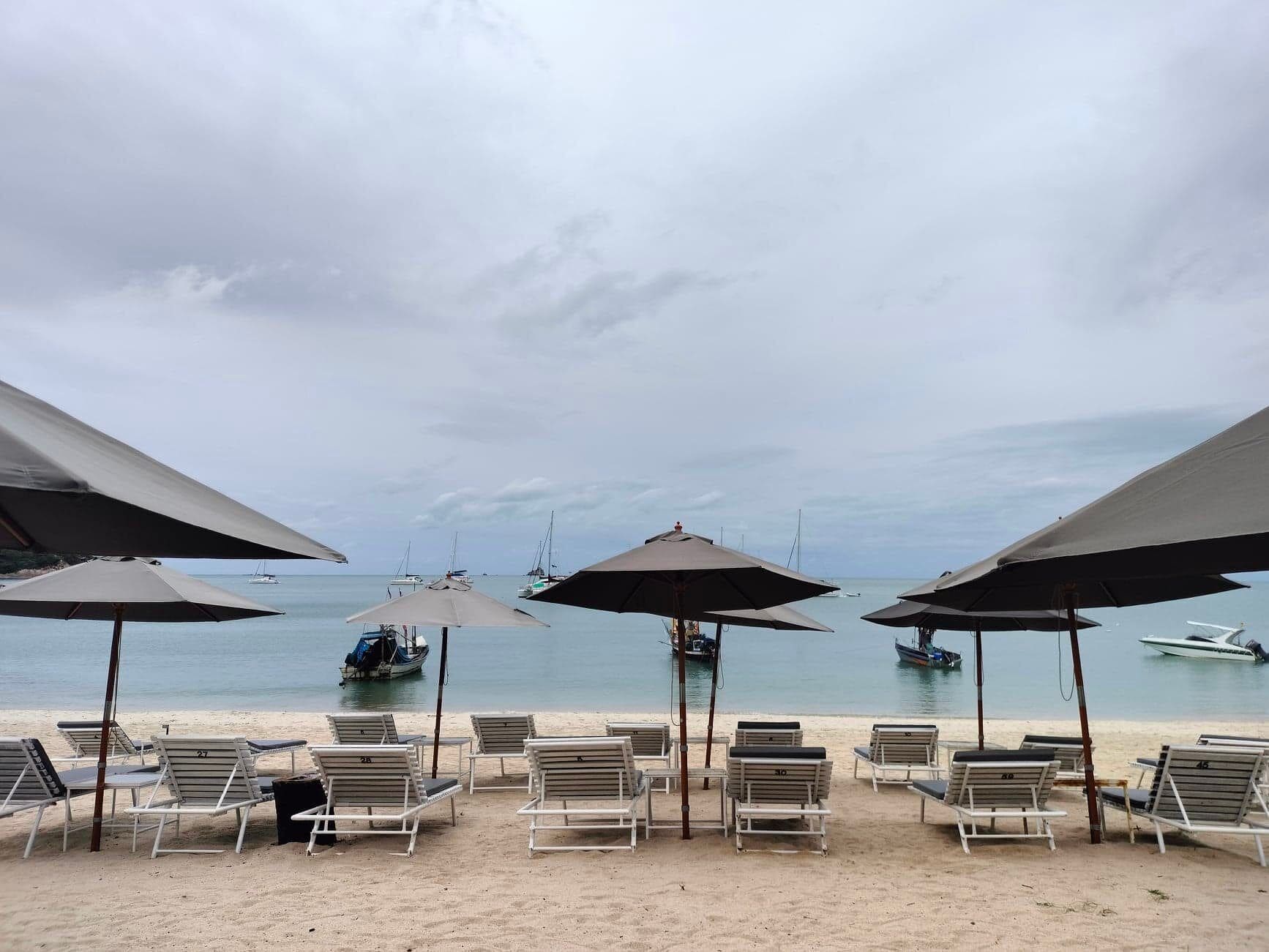 Une plage bordée de rangées de transats et de parasols, des bateaux sur l’eau et un ciel nuageux.