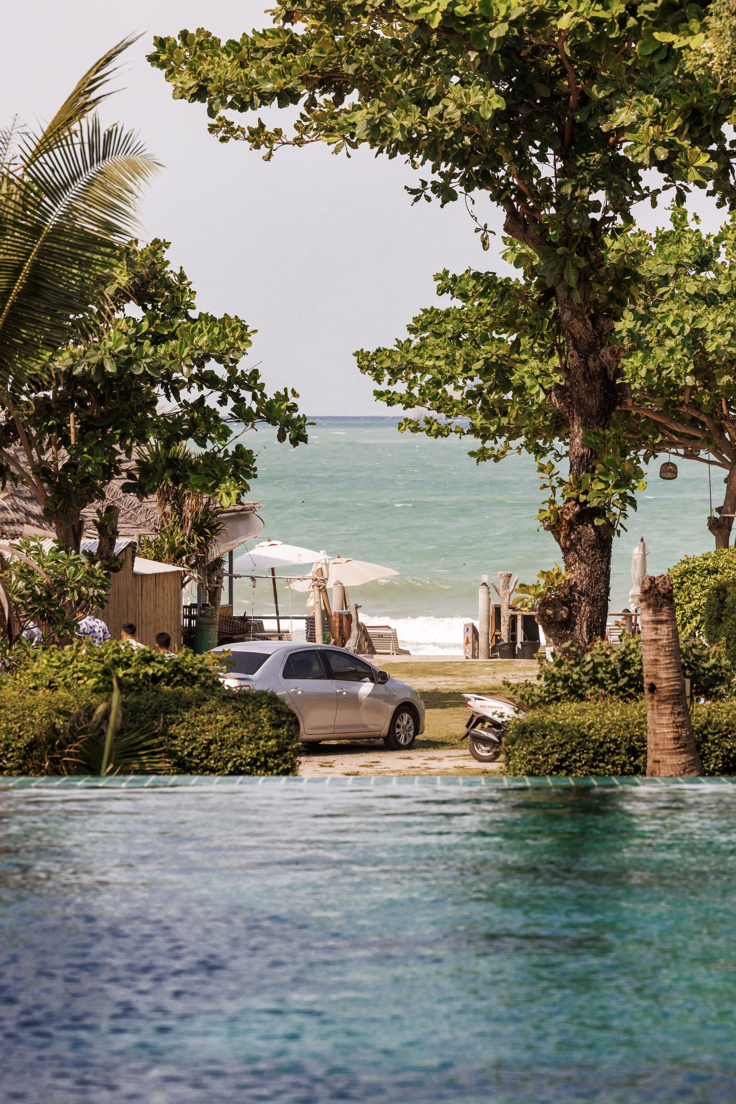 A poolside view with a car parked near the water, surrounded by lush greenery and a beach in the background.