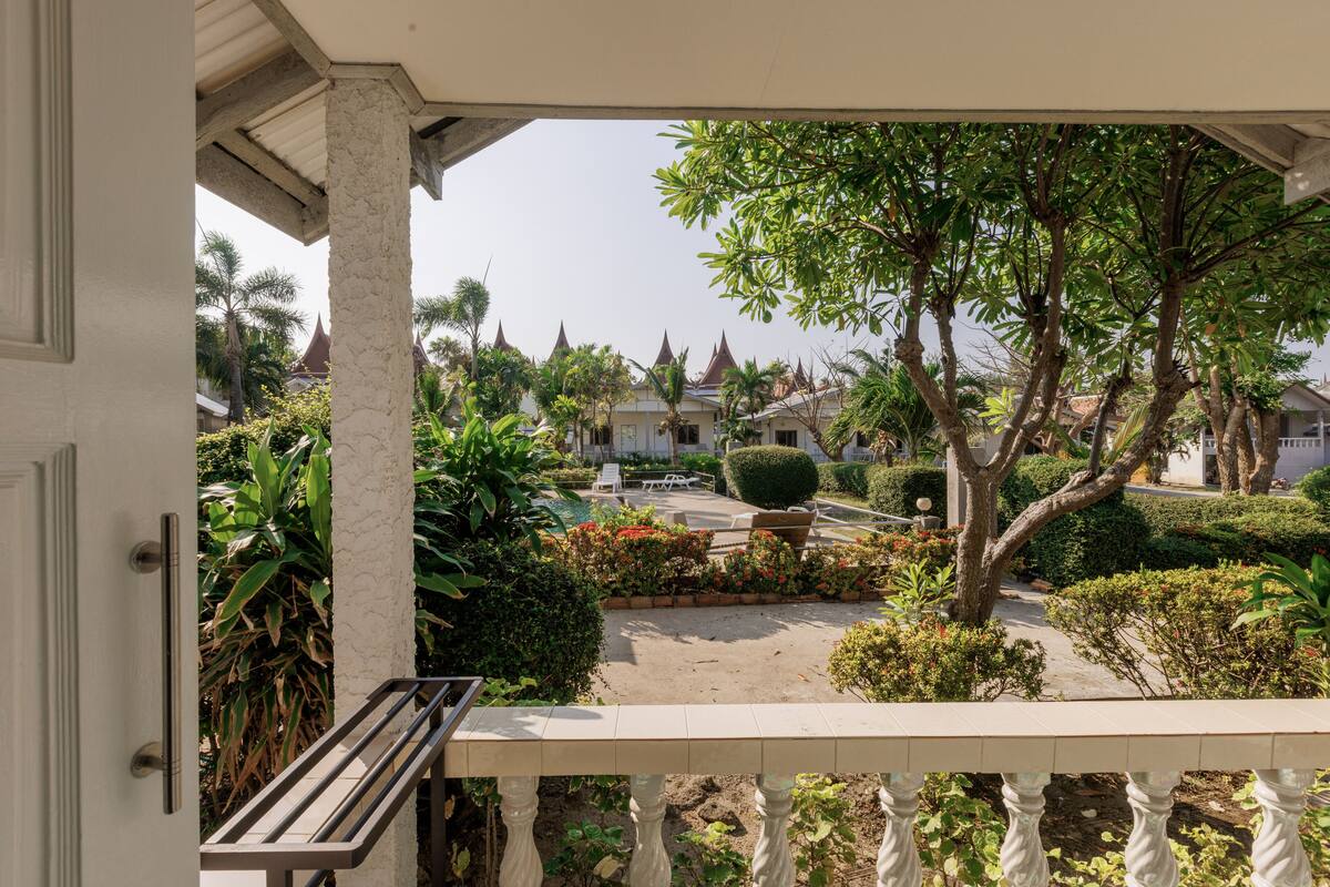 A view from a balcony with a railing, overlooking a garden with various plants and a pathway leading to a building with a traditional roof.