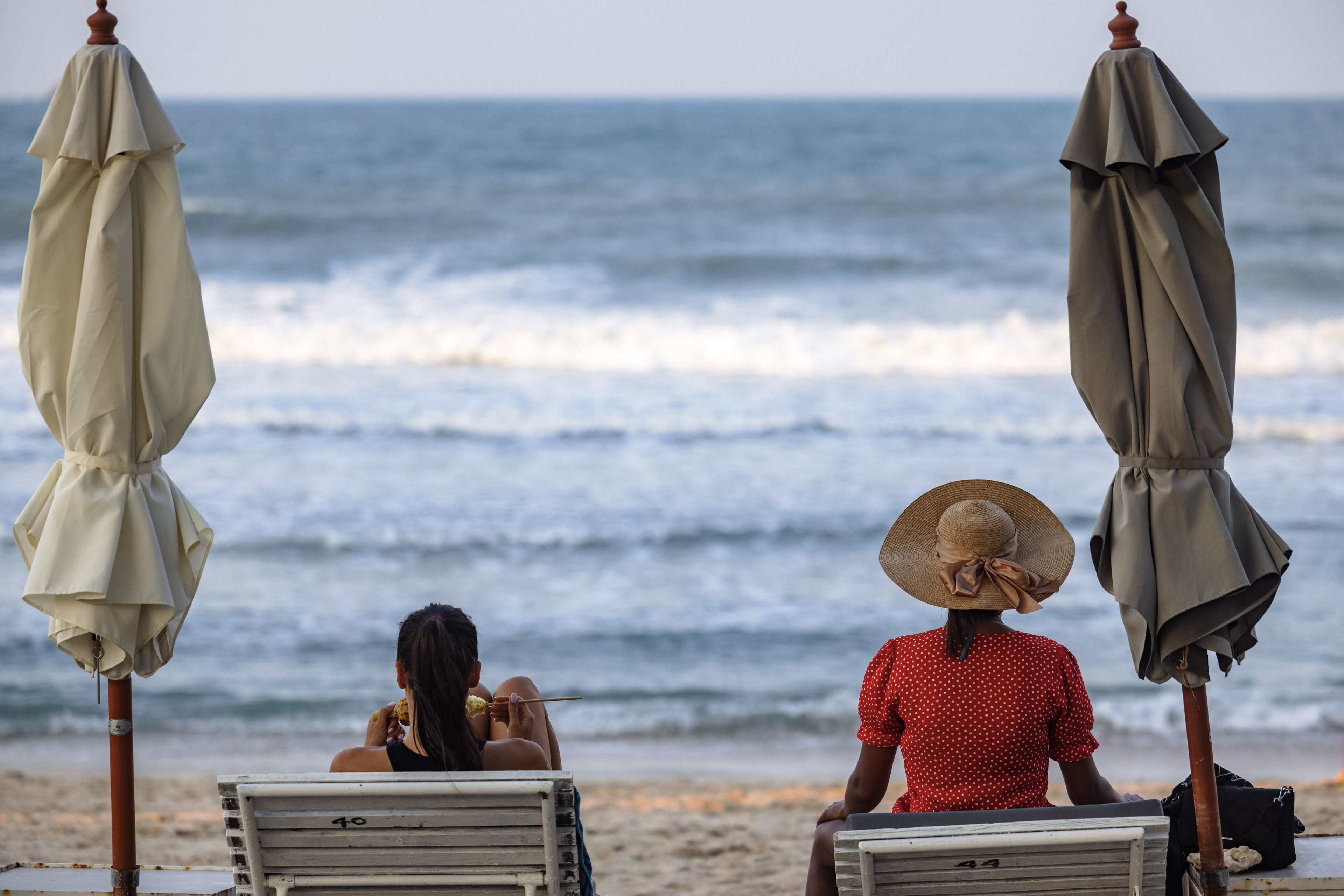 Two beach chairs with umbrellas, a woman in a red polka dot dress and a large hat, and another woman in a black top and white hat.