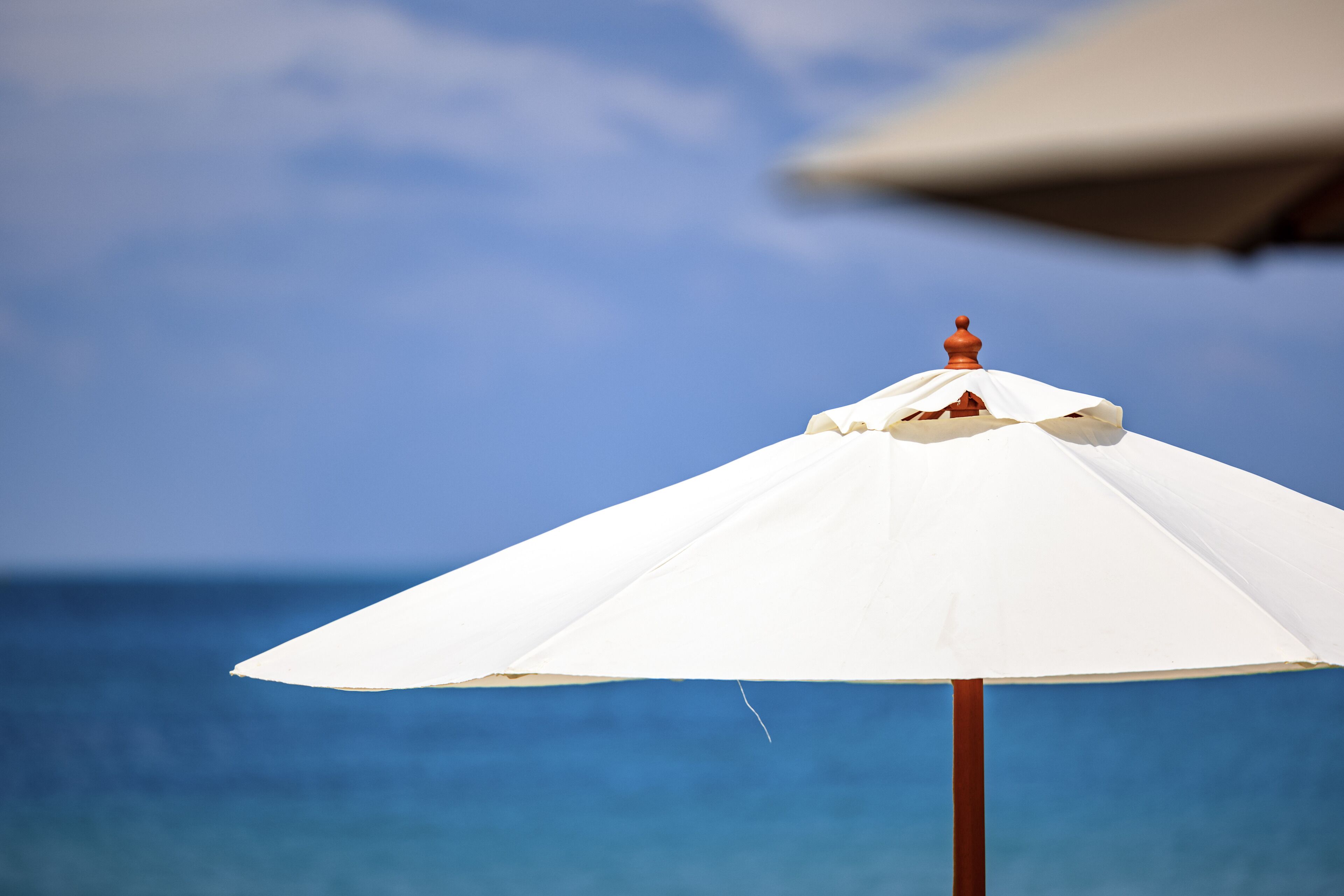 A white beach umbrella with a wooden pole, set against a clear blue sky and ocean.