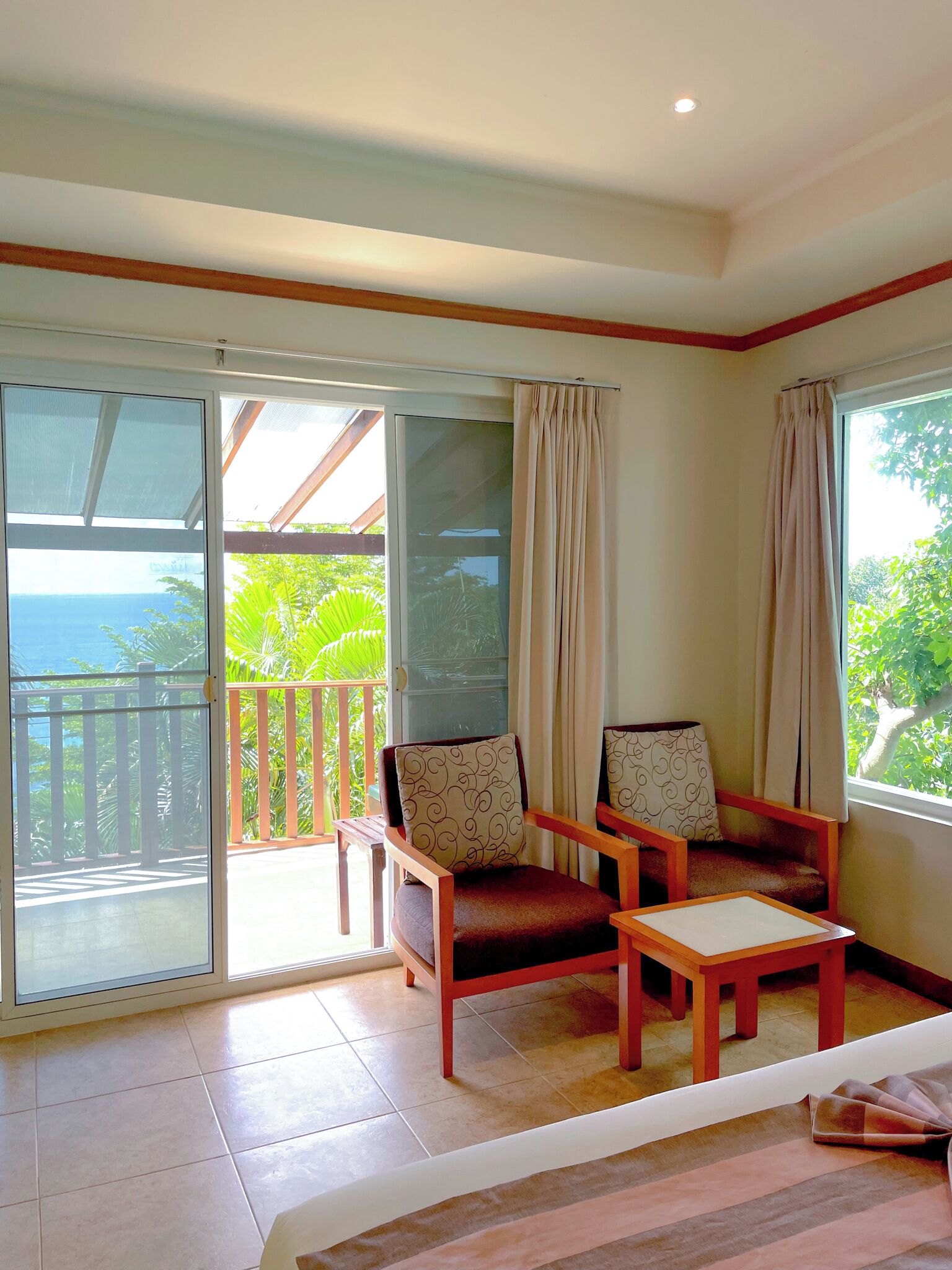 A room with a sliding glass door leading to a balcony with a view of the ocean. There are two wooden chairs with cushions and a small table.