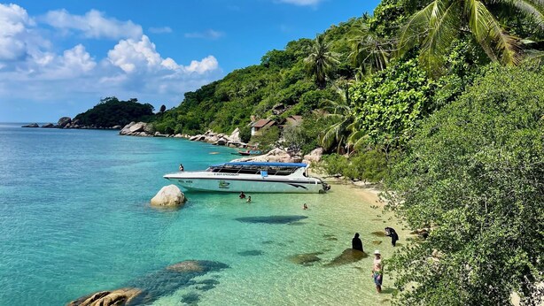 A beach with clear turquoise water, a boat docked near the shore, and lush greenery on the hillside.