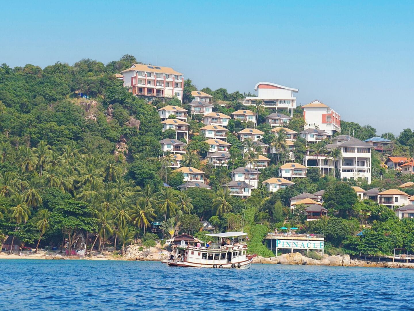 Una zona costera con un barco amarrado cerca de la orilla, rodeada de edificios residenciales y abundante vegetación.