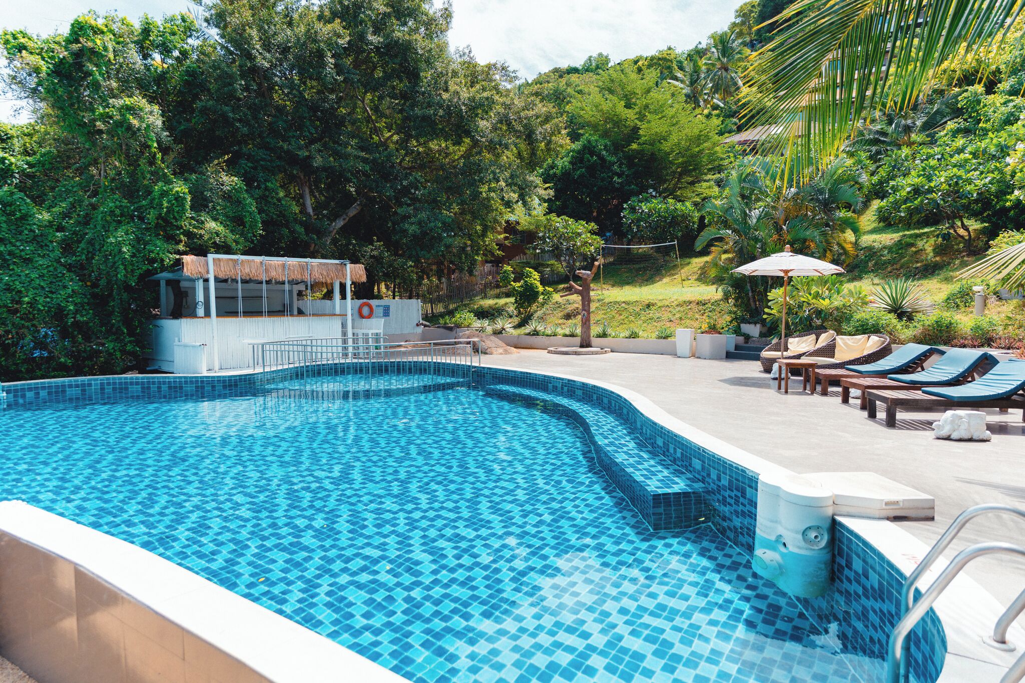 Una piscina con bordes de azulejos, rodeada de sillas de descanso y sombrillas, con una zona a la sombra y vistas a vegetación exuberante.