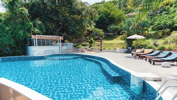 Una piscina con bordes de azulejos, rodeada de sillas de descanso y sombrillas, con una zona a la sombra y vistas a vegetación exuberante.