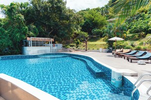 Una piscina con bordes de azulejos, rodeada de sillas de descanso y sombrillas, con una zona a la sombra y vistas a vegetación exuberante.
