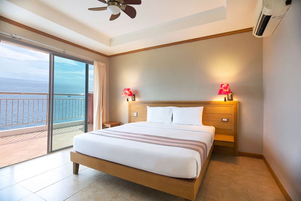 A hotel room with a large bed, two bedside lamps, a ceiling fan, and a view of the sea through a sliding glass door.