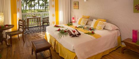 Habitación de hotel con cama, piso de madera, balcón con vista a palmeras y una mesa con sillas.