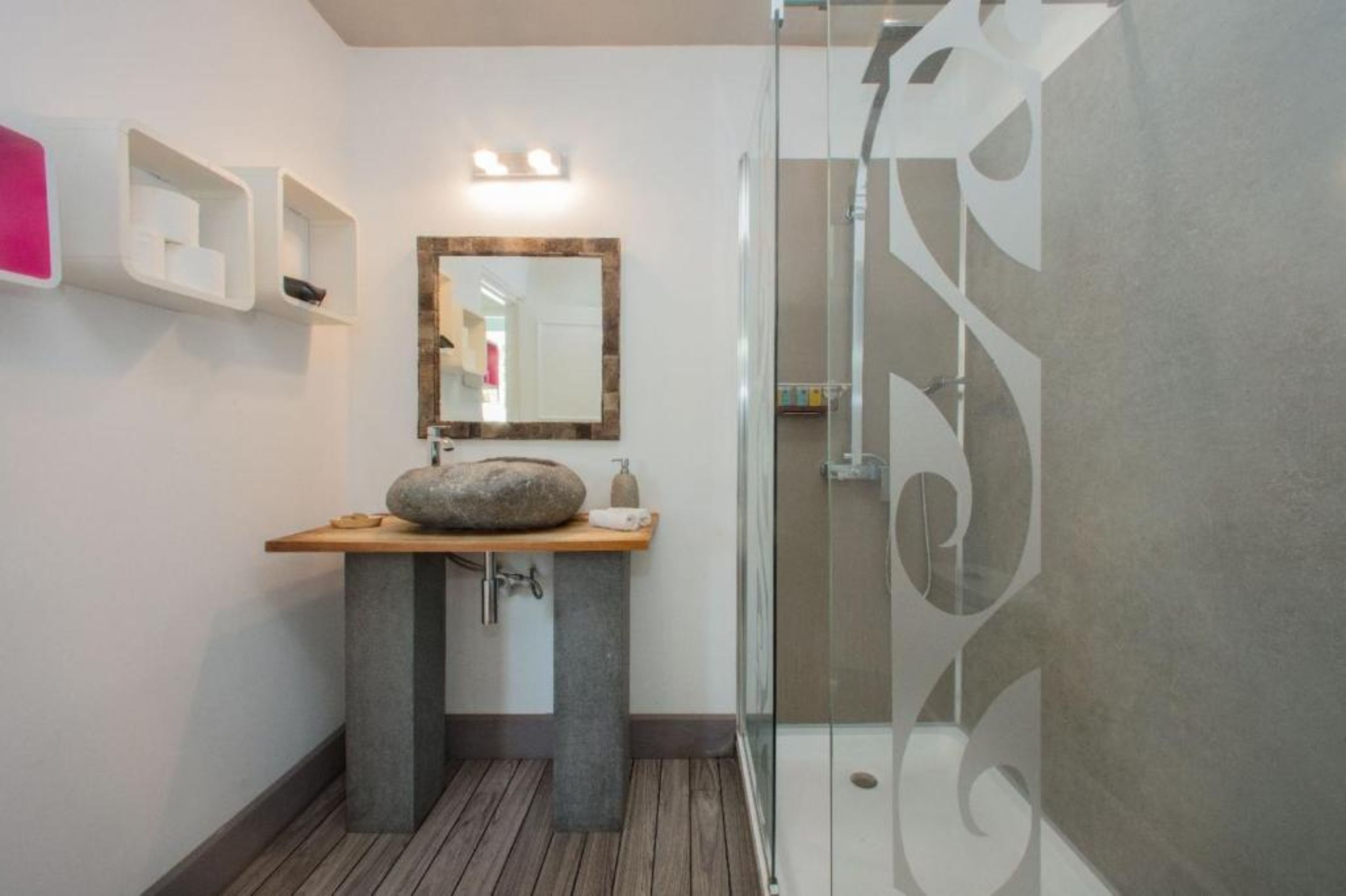 A modern bathroom with a stone sink, a wooden vanity, a glass shower enclosure, and a mirror with a wooden frame.