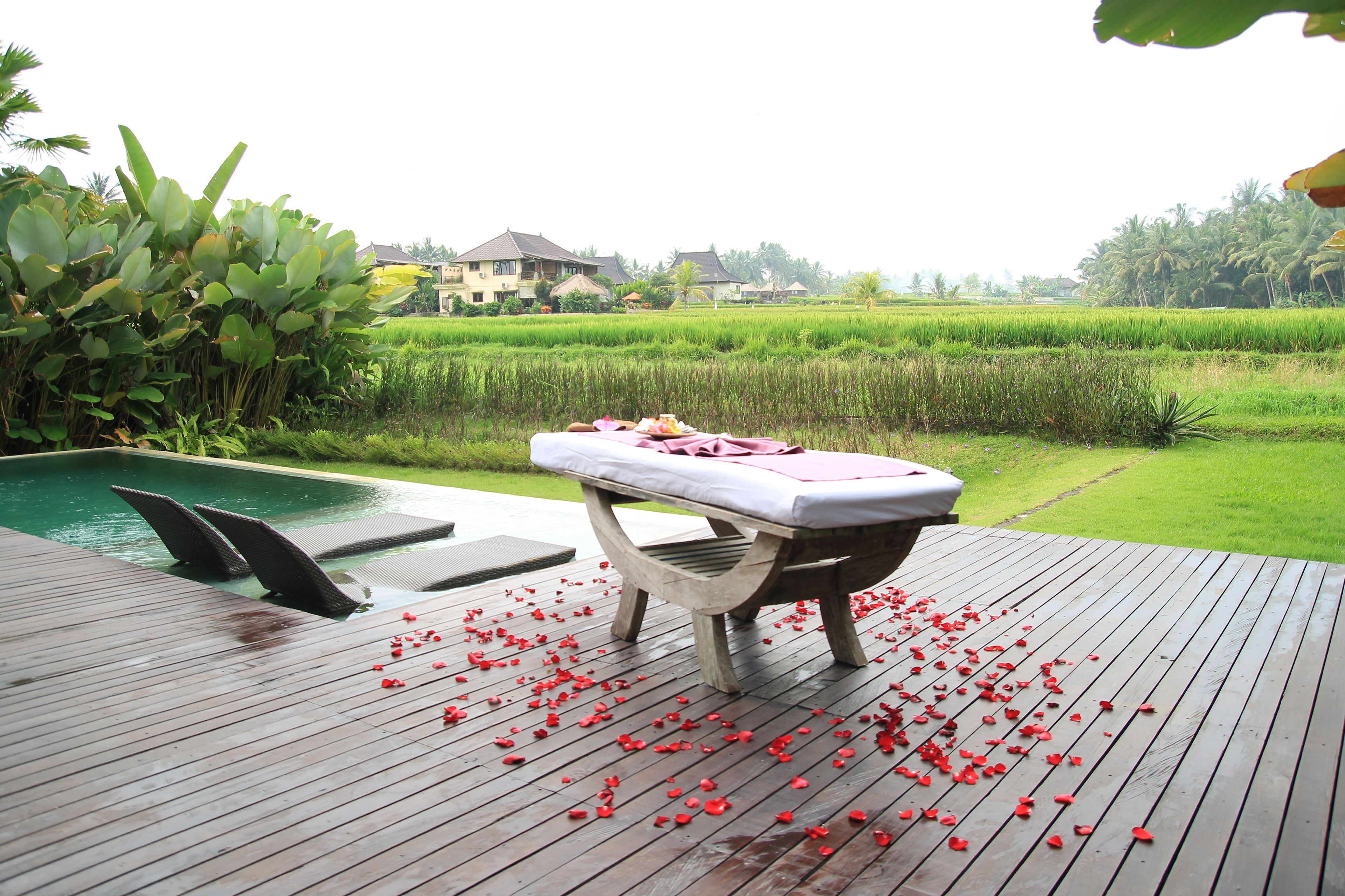 A wooden deck with a massage table and rose petals, overlooking a lush green field and a swimming pool.