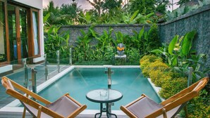 A pool area with two wooden lounge chairs and a small table, surrounded by lush tropical plants and a stone wall. - The Samara (Ubud)