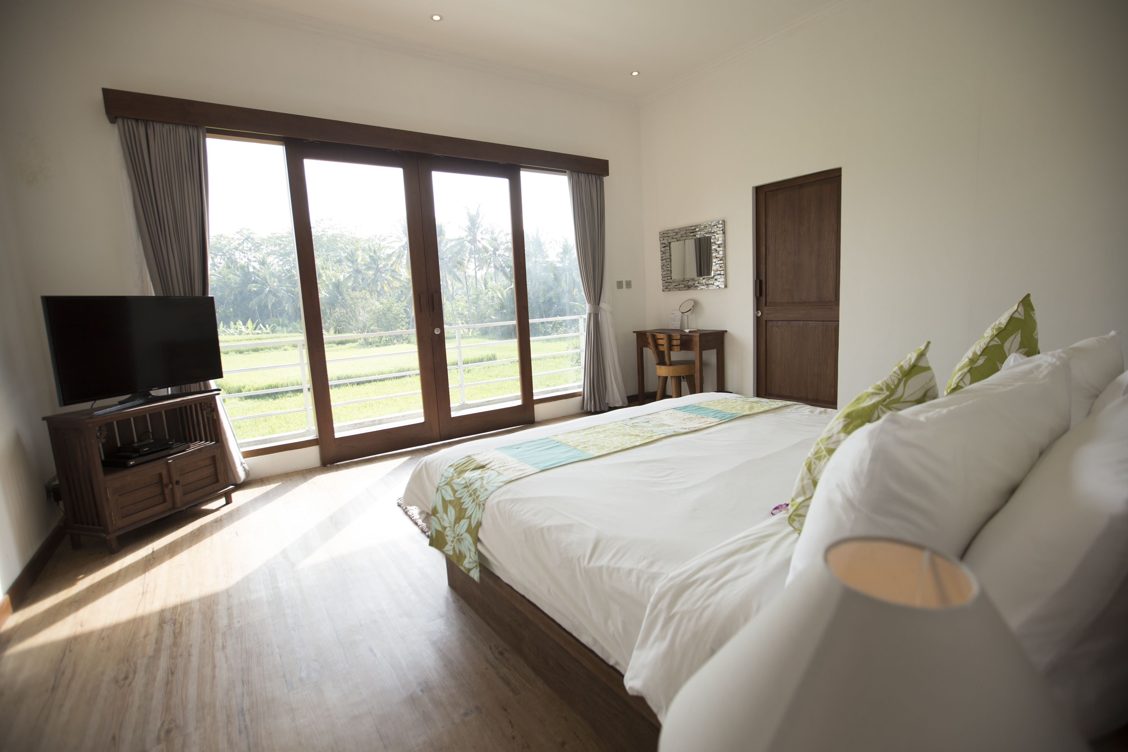 A bedroom with a large bed, a television on a wooden cabinet, a small table with a mirror, and a view of a green landscape through the window.