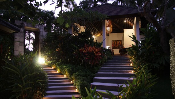 A pathway leading to a building with a thatched roof, surrounded by lush greenery and lit by small lights. - The Samara (Ubud)