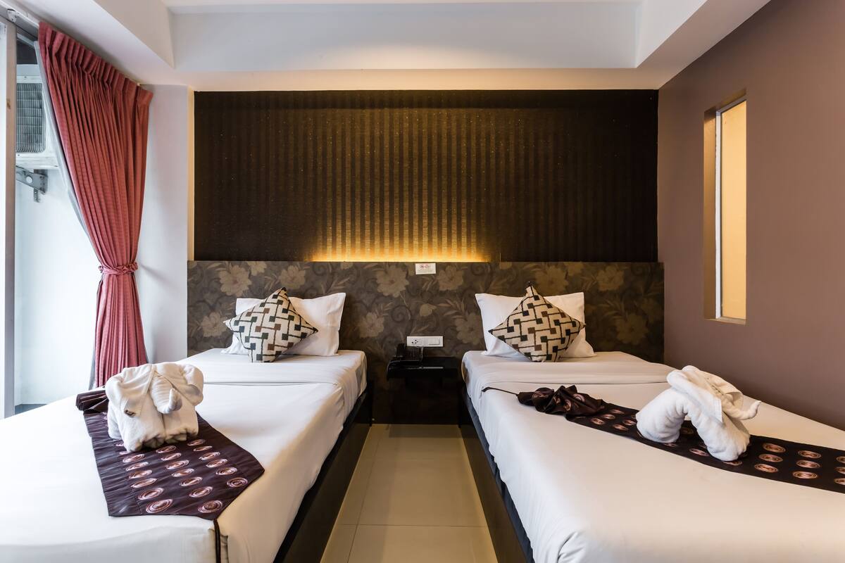 A hotel room with two beds, a large window with a curtain, a wall-mounted light, and a small table with a phone.