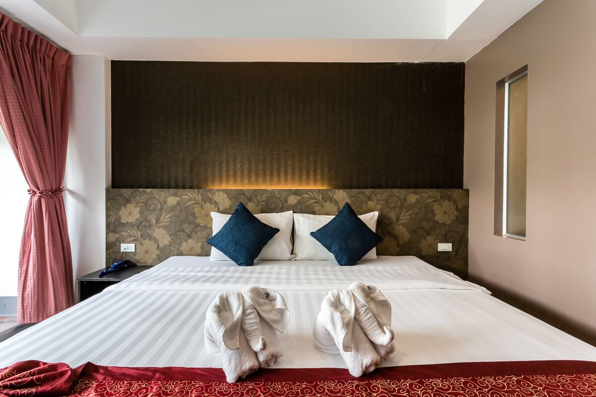 A hotel room with a large bed, two pillows, a nightstand, a red curtain, and a window.