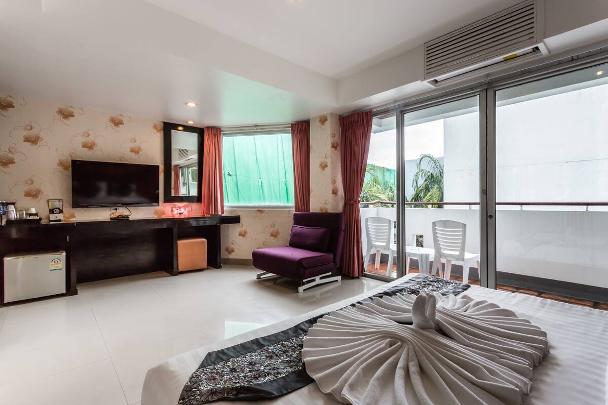 A modern hotel room with a flat-screen TV, a small refrigerator, a desk with a computer, a purple chair, and a balcony with white chairs.