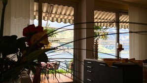 A room with a view of a mountainous landscape through large windows. There is a dining area with a table and chairs visible through the window. - Villa Edera (Malcesine)