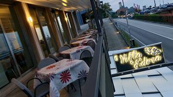 An outdoor dining area with tables covered in floral tablecloths, a sign reading "Villa Edera," and a view of a road with parked cars and a castle-like structure in the background.