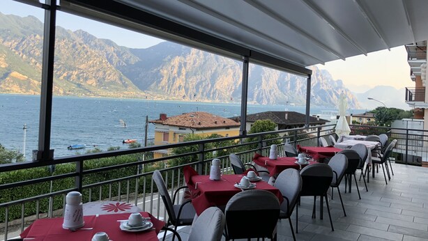 A terrace with tables set for dining, overlooking a lake with boats and mountains in the background. - Villa Edera (Malcesine)