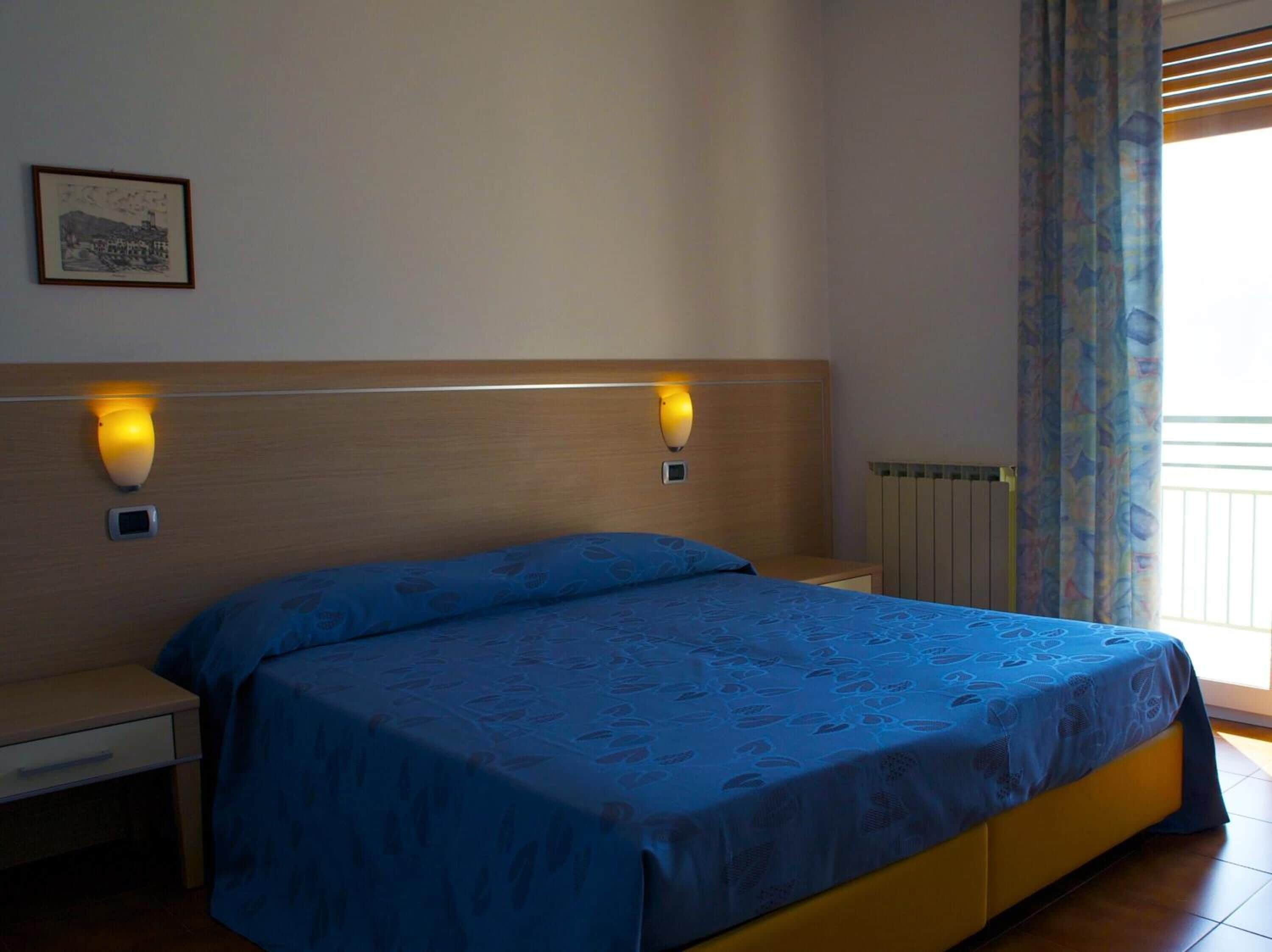 A bedroom with a bed, two bedside tables, a nightstand, a wall-mounted light, a framed picture, and a window with curtains.