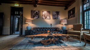 A cozy room with a leather sofa, a wooden coffee table, and two lamps. There are framed artworks on the wall and a window with a view. - Fletcher Landgoedhotel Renesse (Renesse)