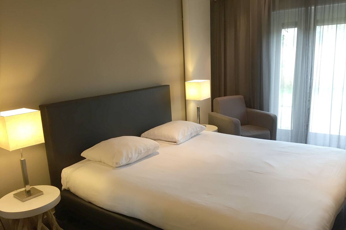 Habitació d'hotel amb un llit gran, dues làmpades de nit, una cadira i una finestra amb cortines.