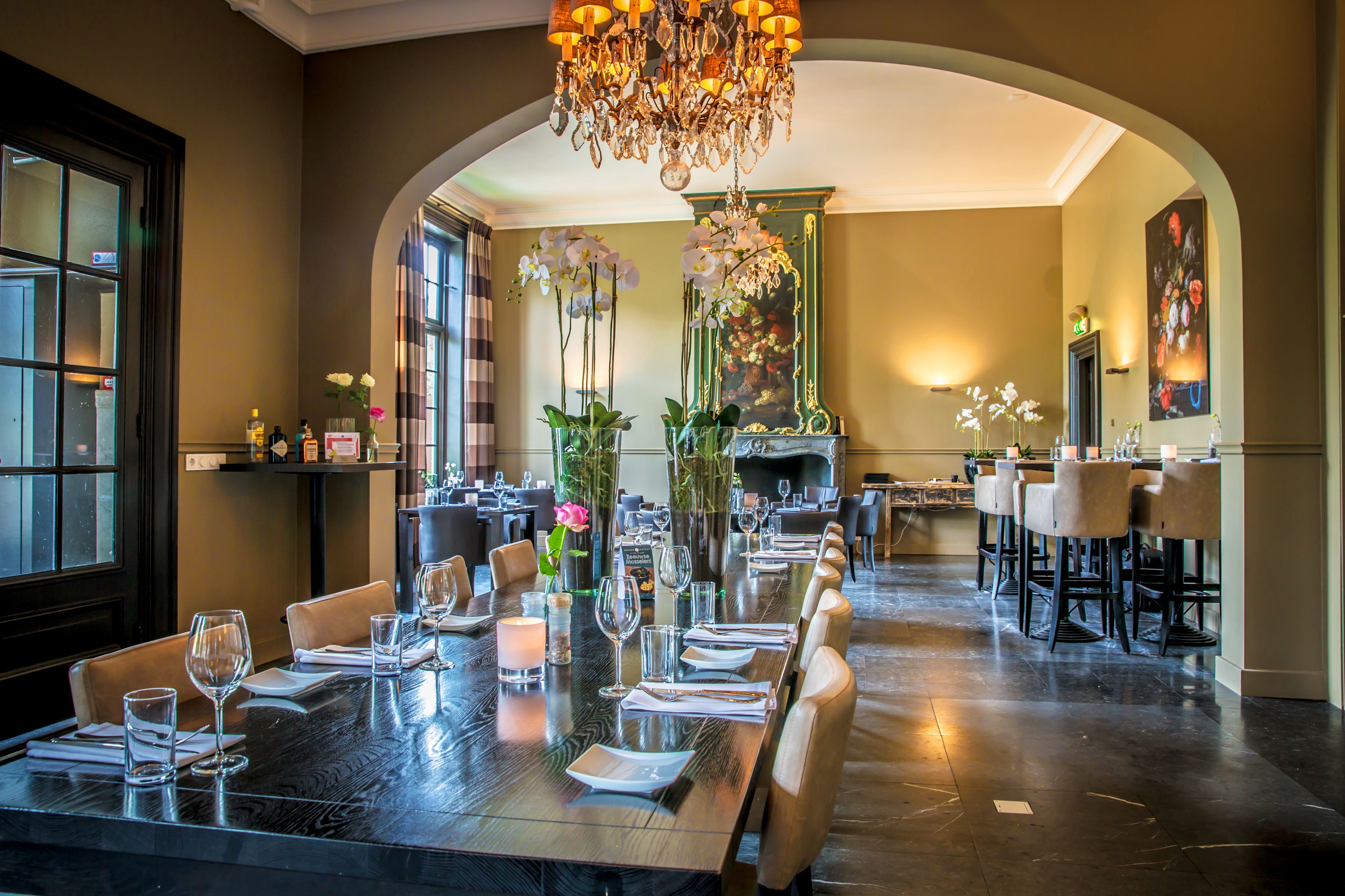A dining area with a long table set for a meal, surrounded by chairs, with a chandelier overhead and a large window on one side.