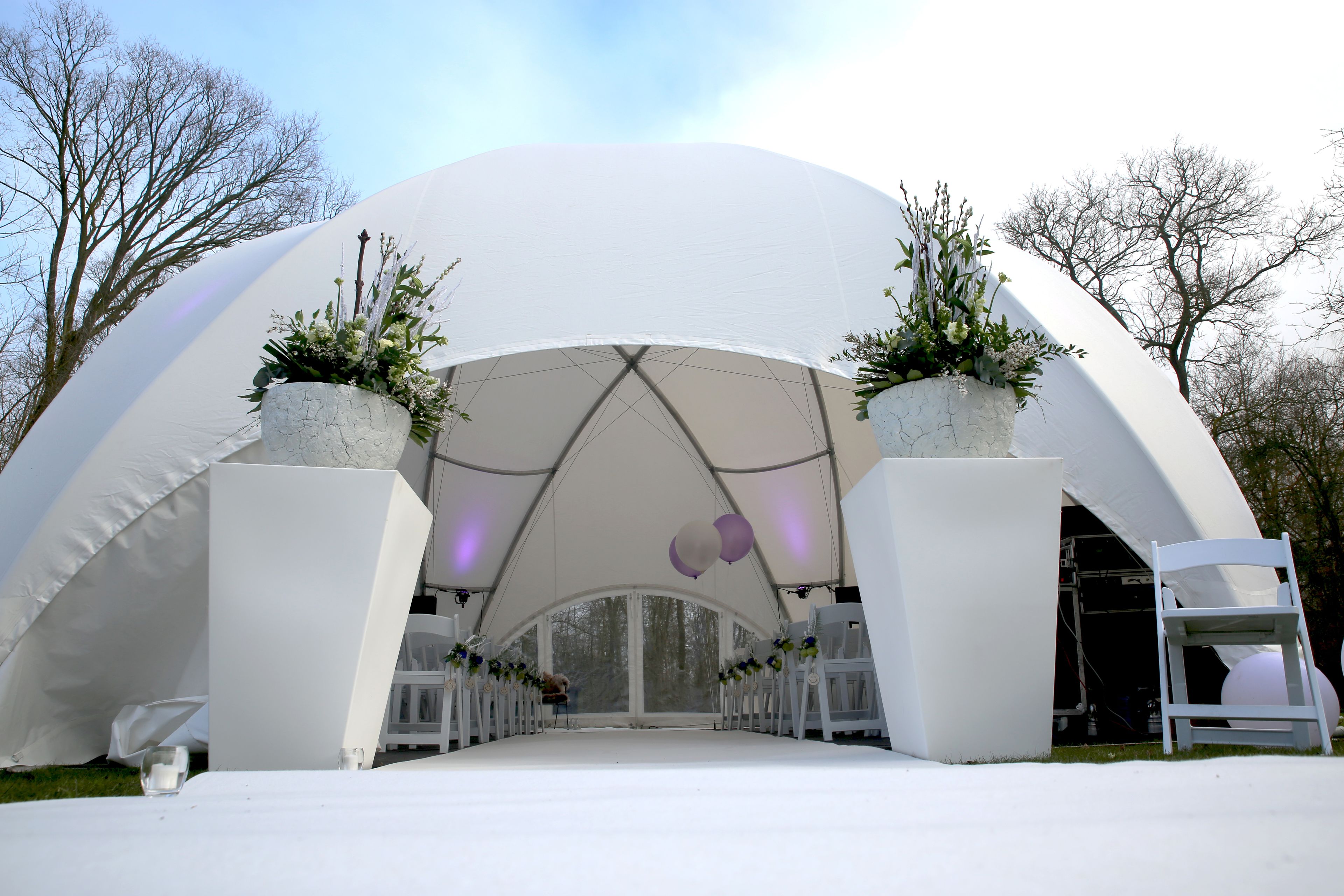 A large white tent with a semi-circular archway, surrounded by white chairs and potted plants.