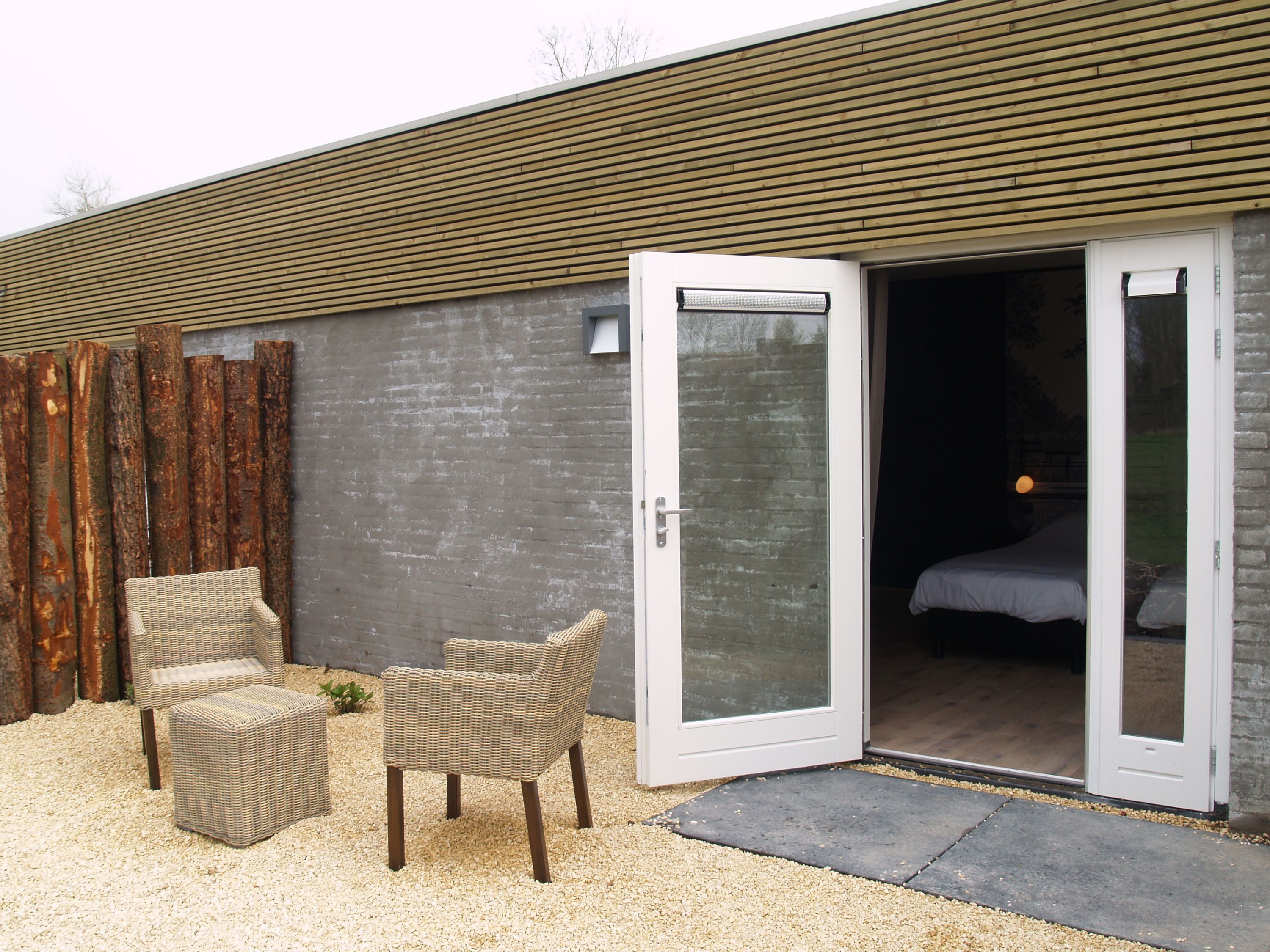 A modern building with a sliding glass door leading to a bedroom. There are wicker chairs outside and a gravel ground.