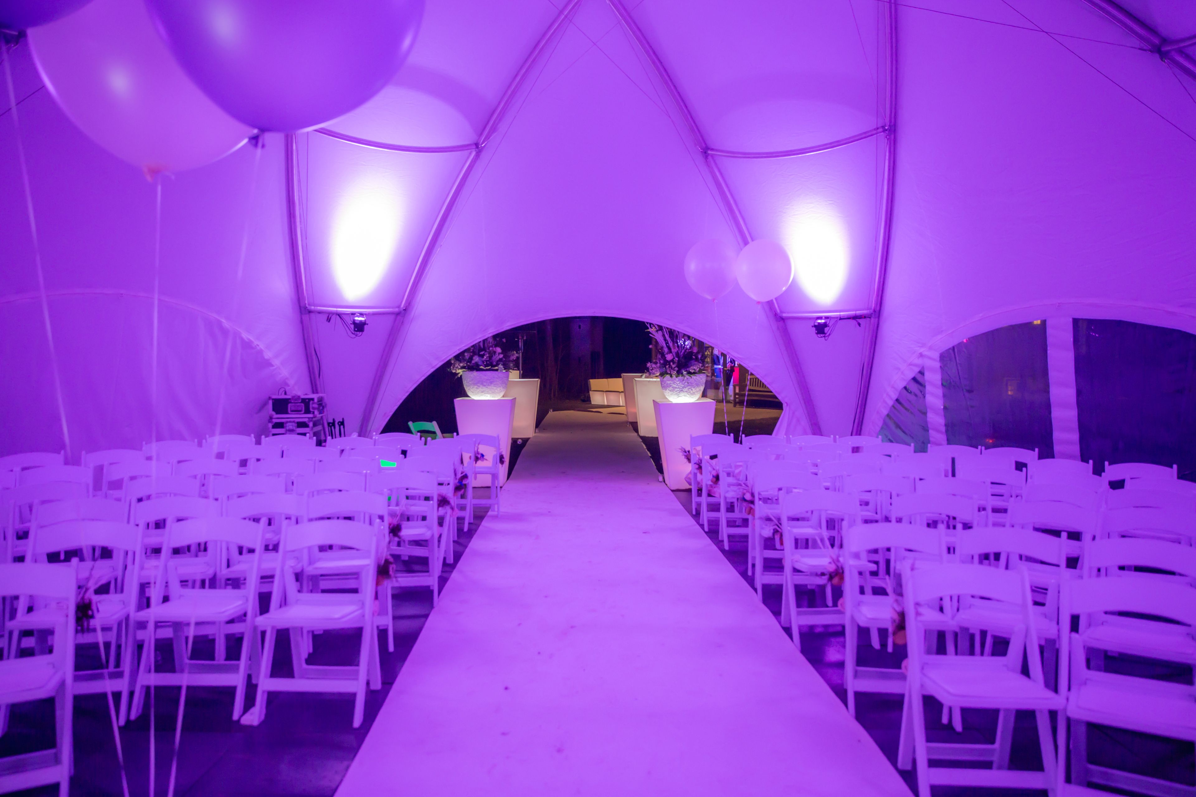 Un espacio interior para eventos con pasillo alfombrado en blanco, sillas blancas a ambos lados y un pasillo central que conduce a una zona de escenario.