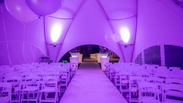 Un espacio interior para eventos con pasillo alfombrado en blanco, sillas blancas a ambos lados y un pasillo central que conduce a una zona de escenario.